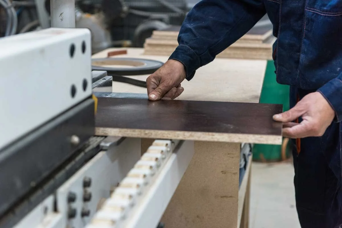 A person working in a woodworking shop, handling a dark wood veneer sheet at a machine.