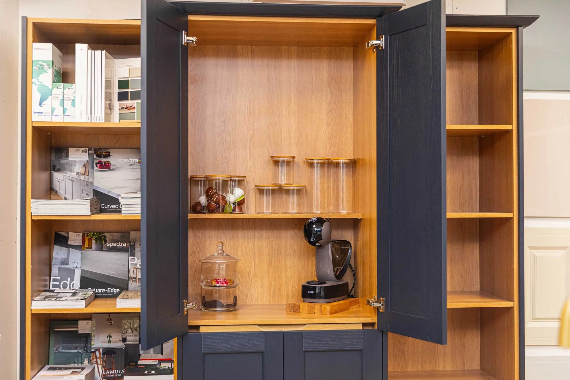 Empty wooden cabinet with black doors, shelves holding glass jars, a coffee maker, and a dish with a treat, surrounded by other similar shelves with magazines and books.