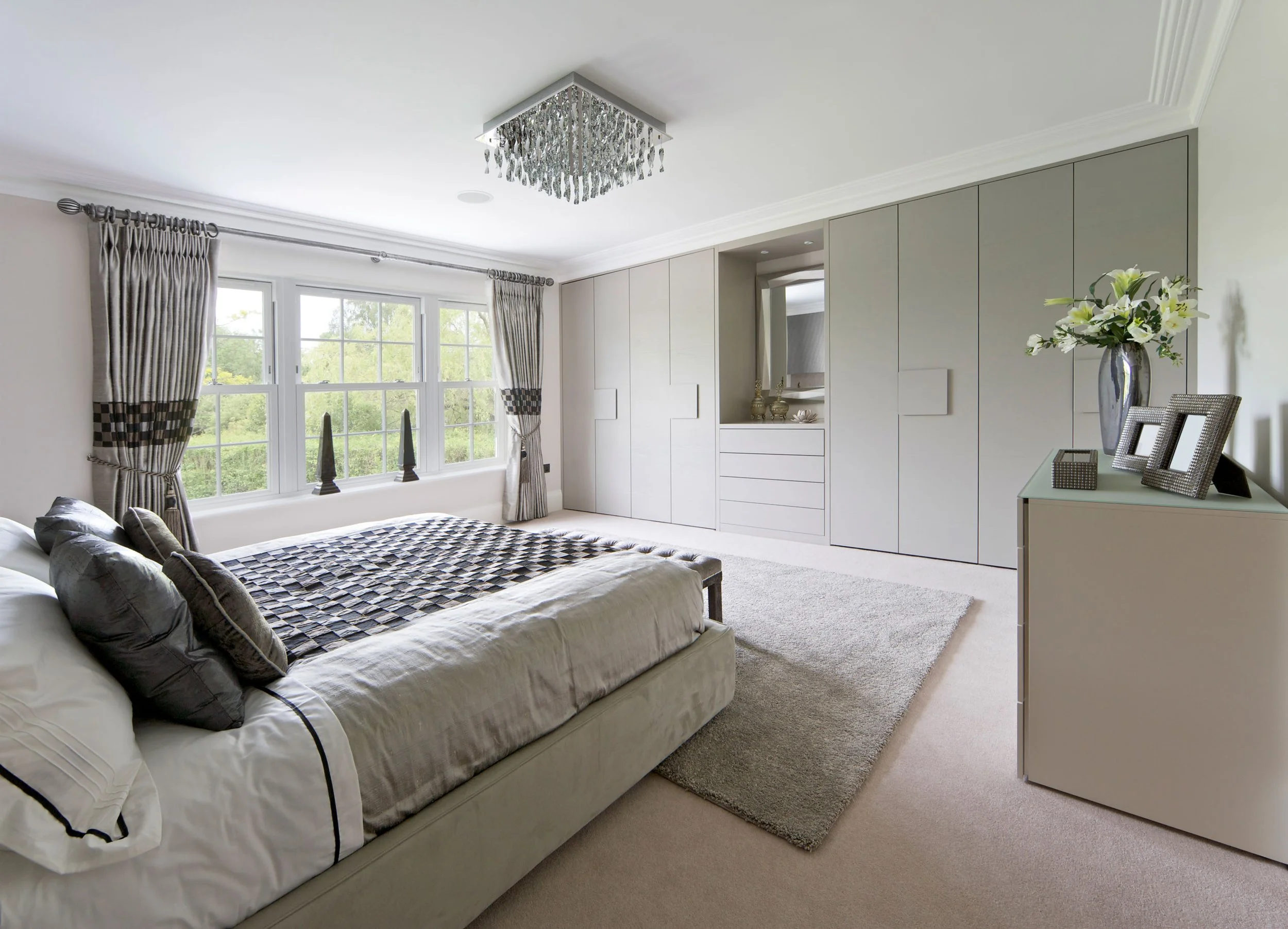A bedroom with a large window dressed with curtains, a bed with gray and white bedding, a beige carpet, and a built-in closet with drawers and decorative items. A chandelier hangs from the ceiling and there is a side table with a vase of flowers and picture frames.