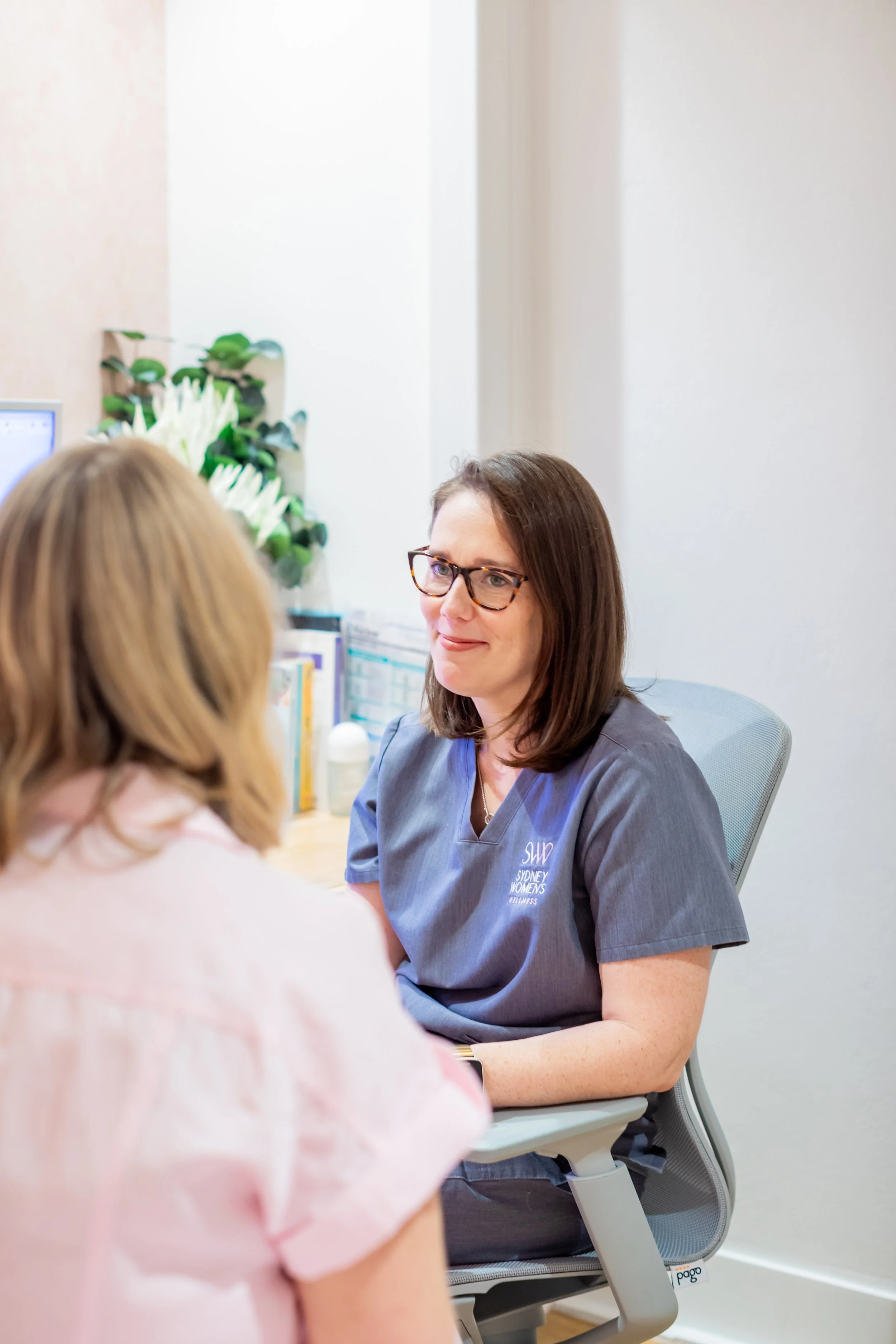 A woman wearing glasses and a gray uniform is sitting at a desk, engaging in conversation with another woman who has blonde hair. The setting appears to be a professional office or clinic with a computer, books, and a flower arrangement in the background.