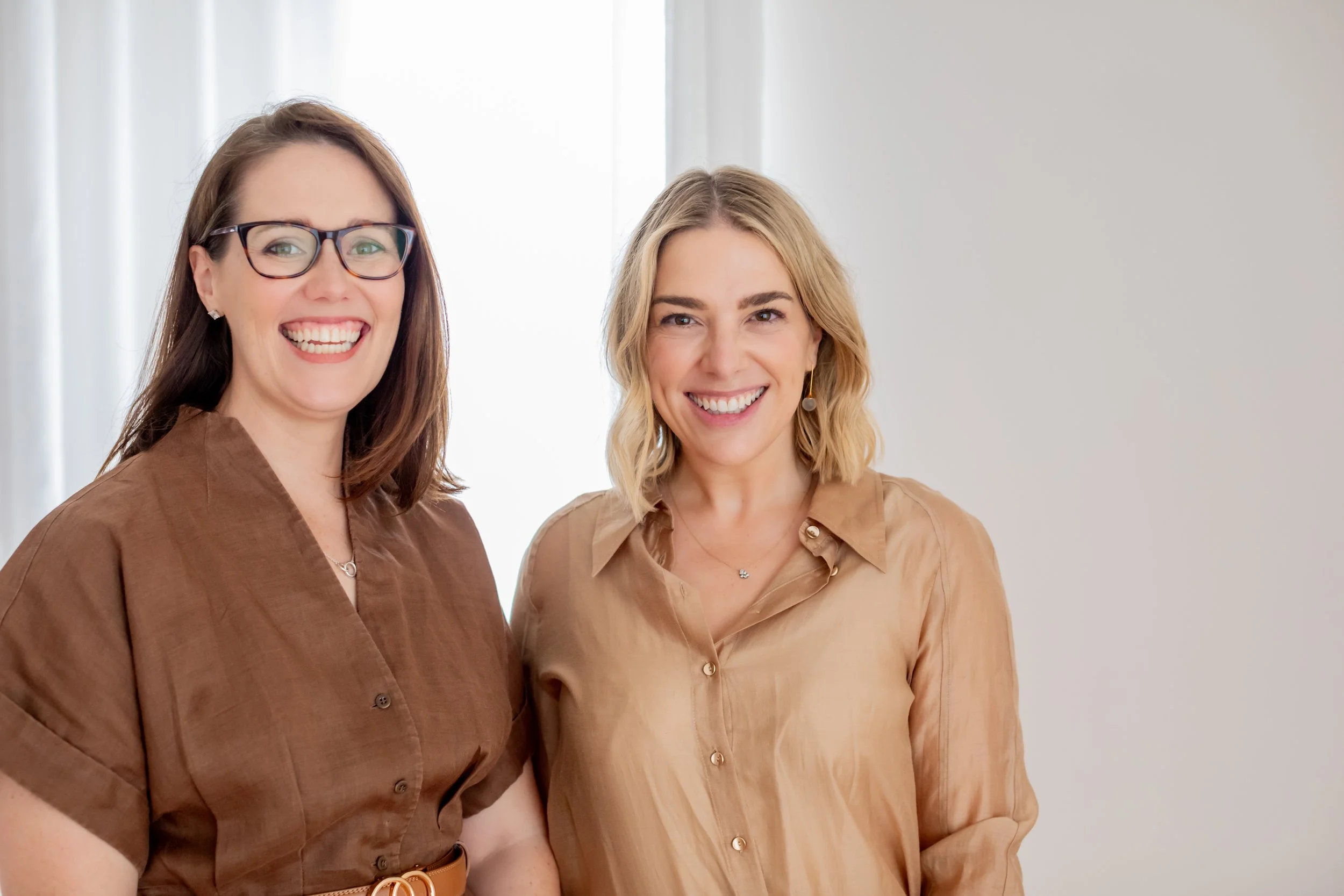 Two women smiling, standing close together in front of a light-colored wall and window with white curtains.