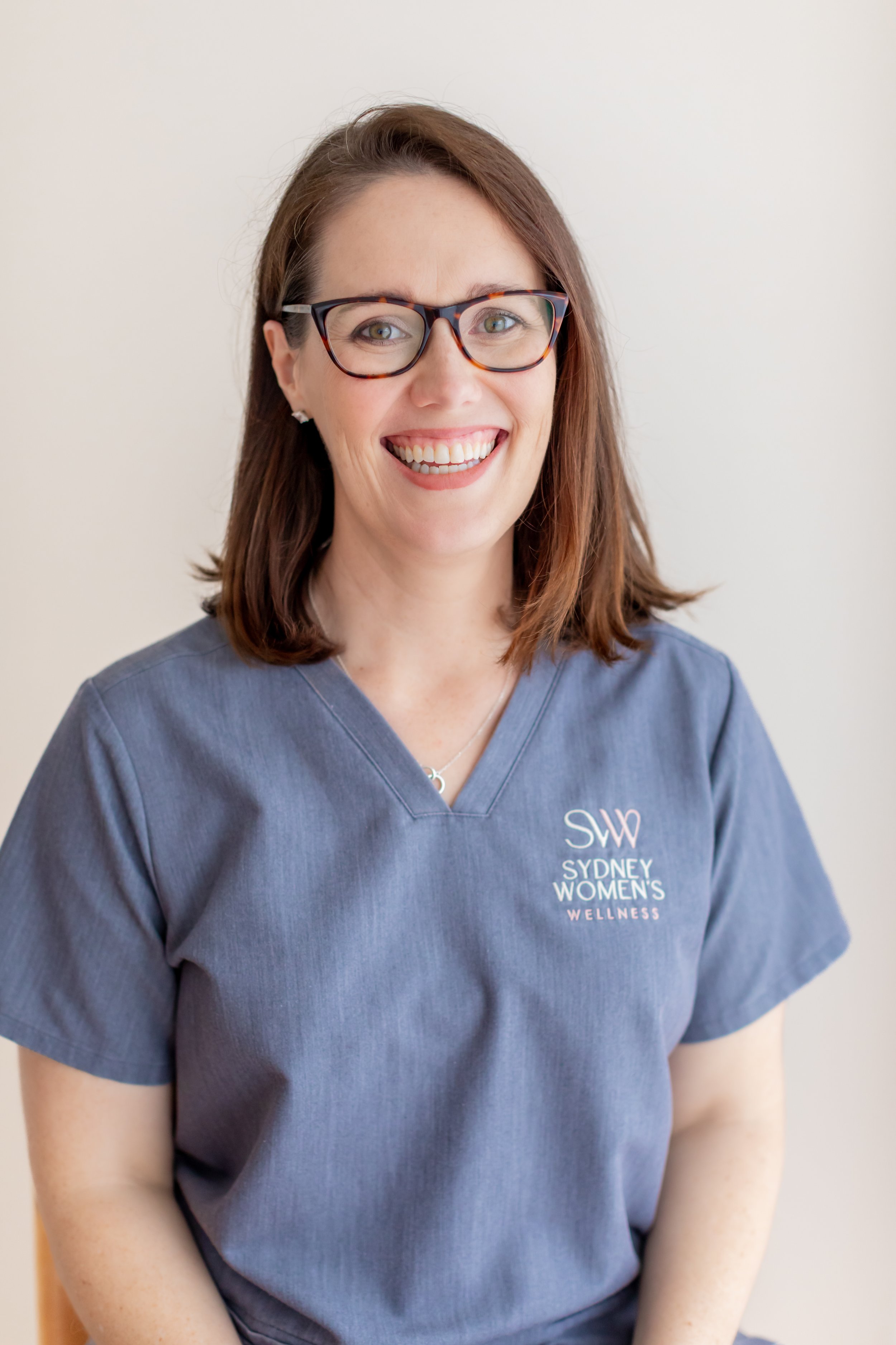A woman with brown hair, glasses, and a big smile, wearing a blue scrub top with the logo 'Sydney Women's Wellness'