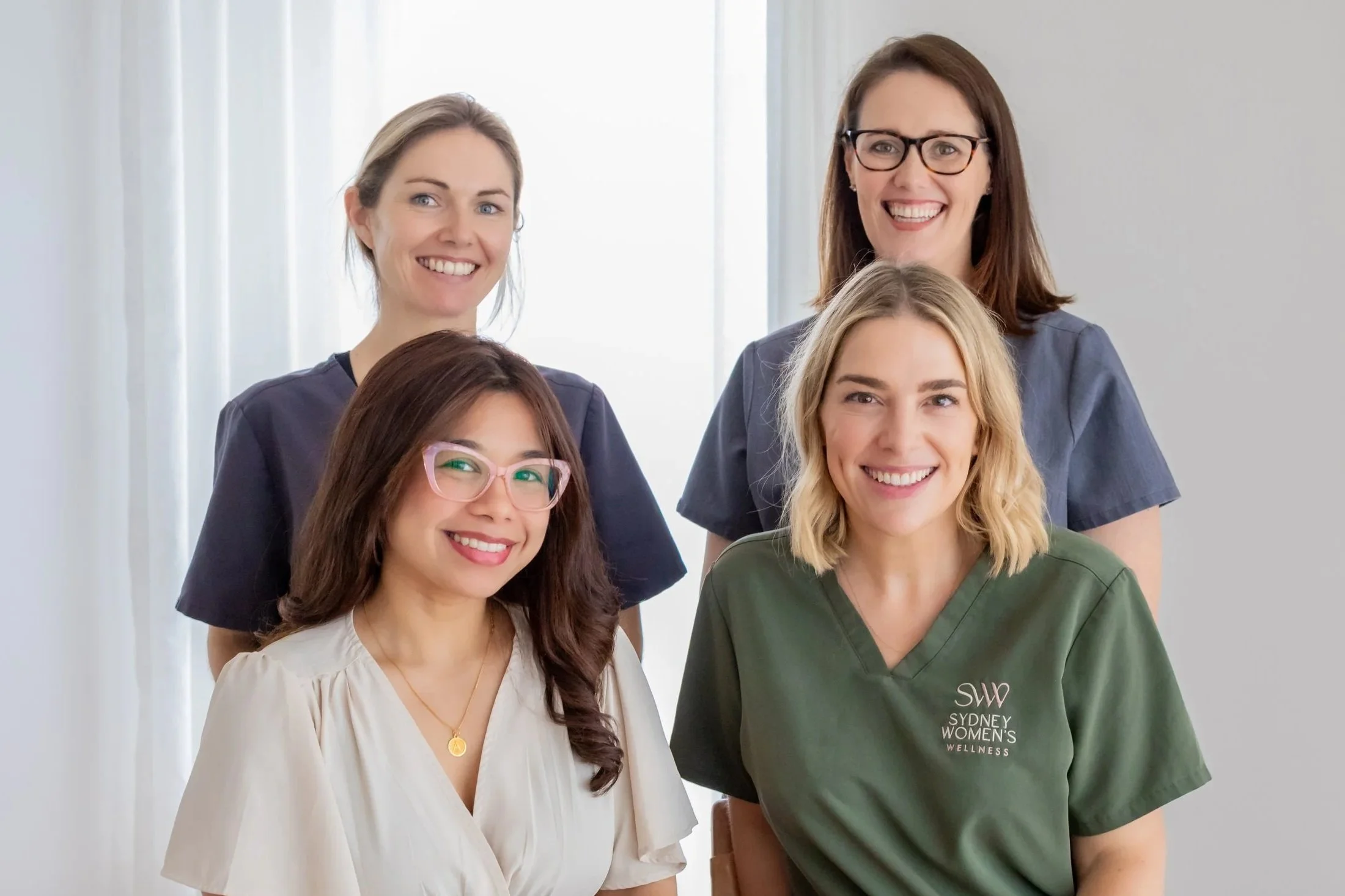 Group of five women, three standing and two seated, smiling indoors with light-colored curtains in the background. One woman is wearing glasses, and one has a badge with 'SW Sydney Women's Wellness' on her scrubs.