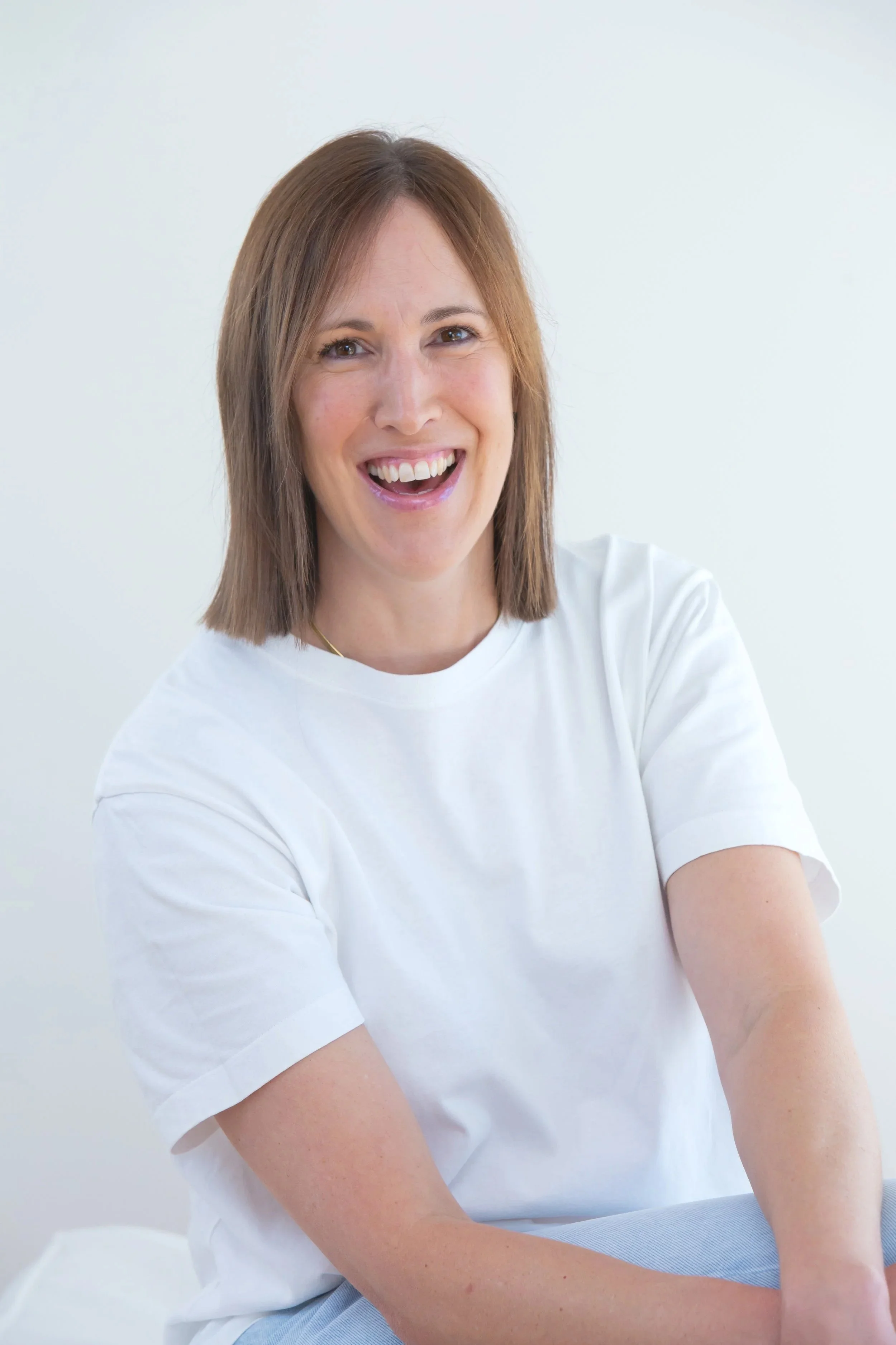 A woman smiling with braces on her teeth, wearing a white t-shirt, sitting against a plain white background.