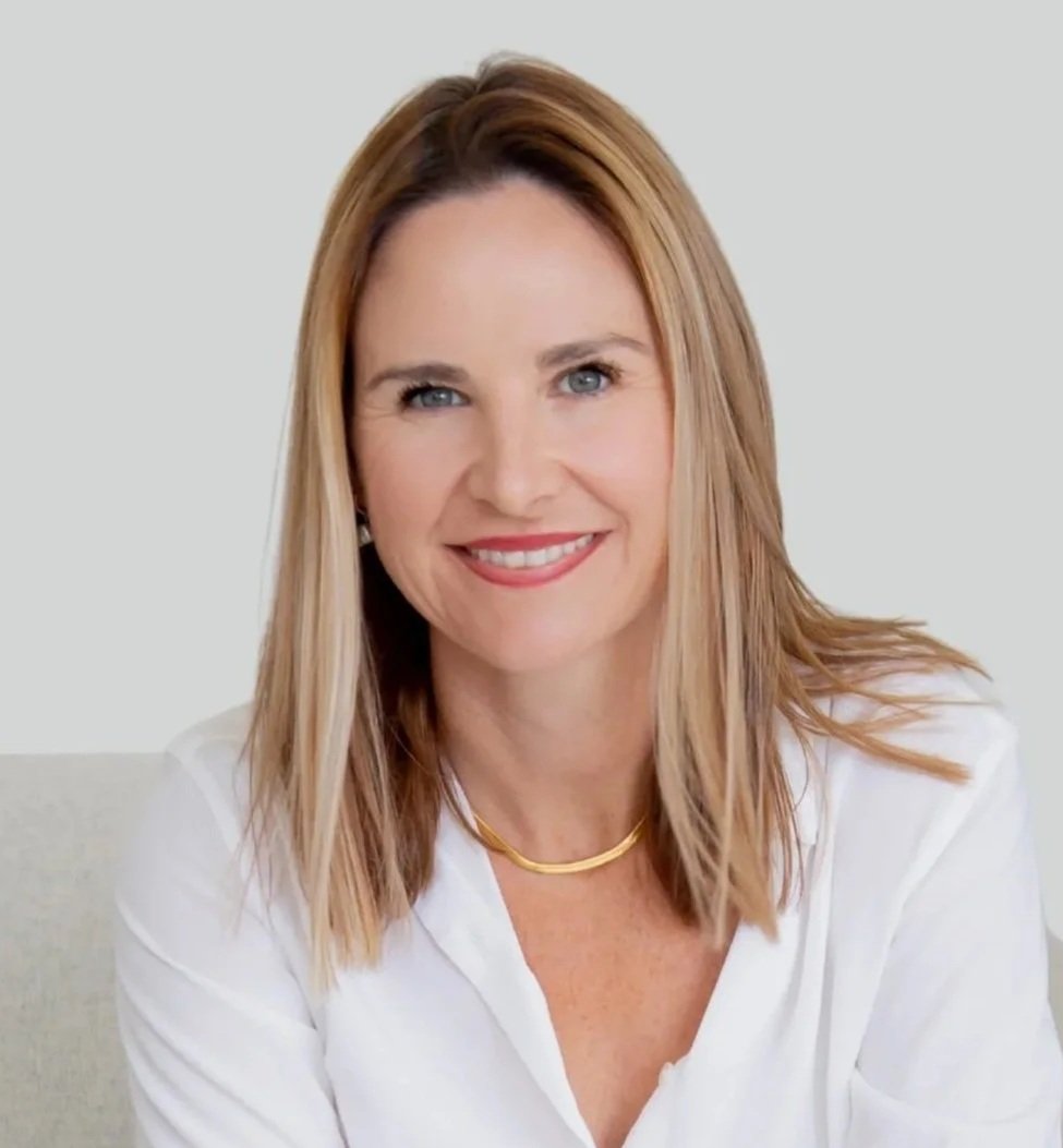 Portrait of a smiling woman with blonde hair wearing a white blouse and a gold necklace, sitting against a plain background.