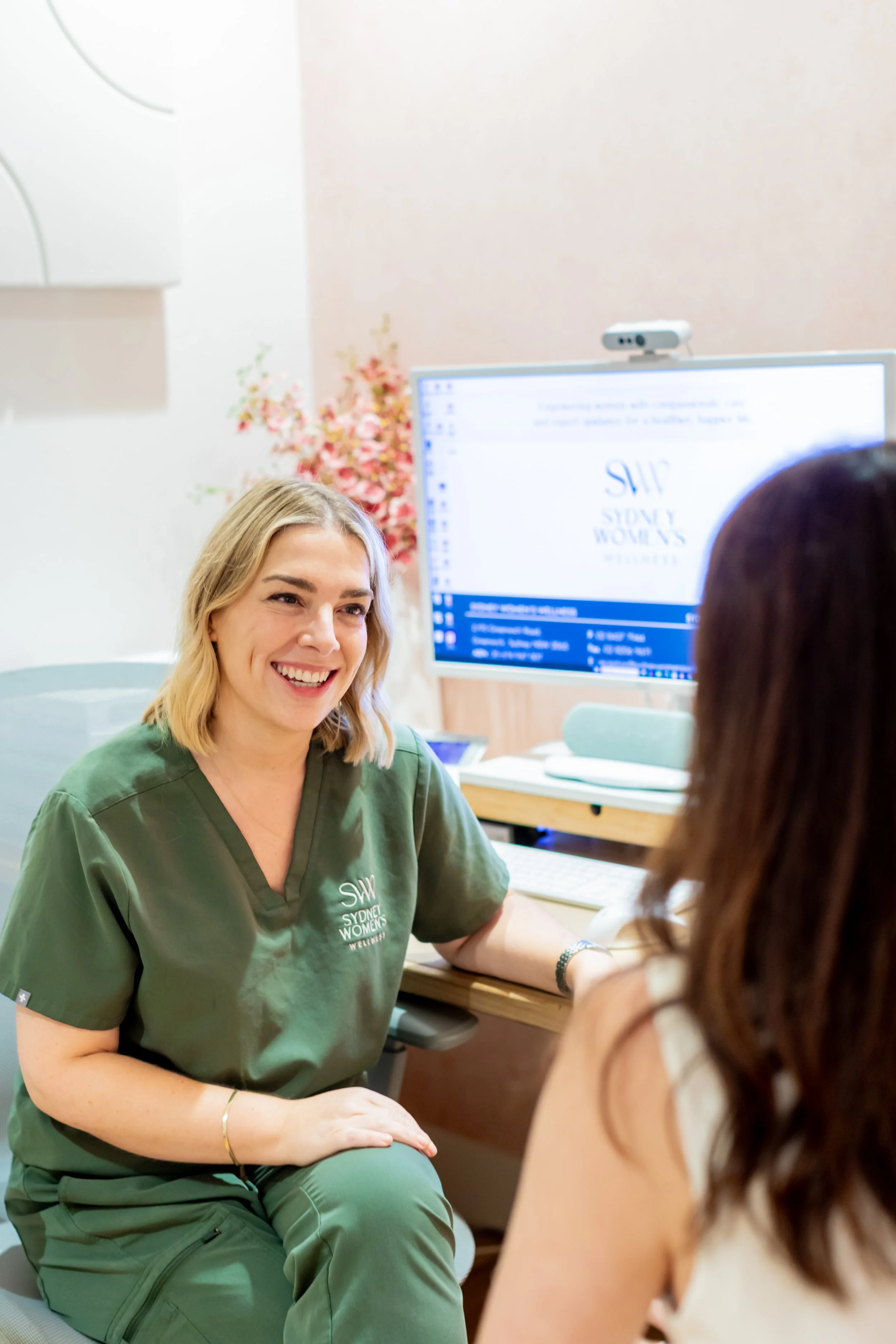 A woman with blonde hair wearing a green uniform smiling and talking to another woman with long brown hair in a white top at a medical office table. Behind them is a computer monitor and a pink flower arrangement.