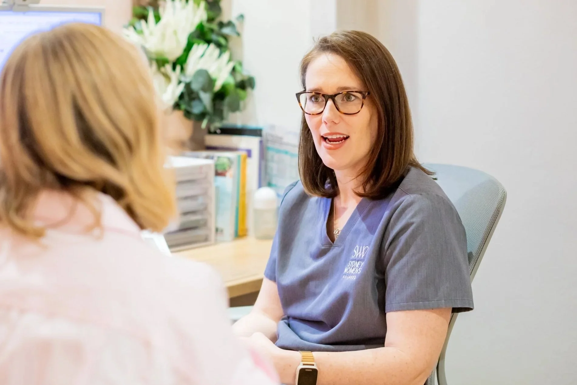 A woman with dark hair, glasses, and a gray medical scrub top sitting at a desk, having a conversation with another person, with a medical office setting in the background.