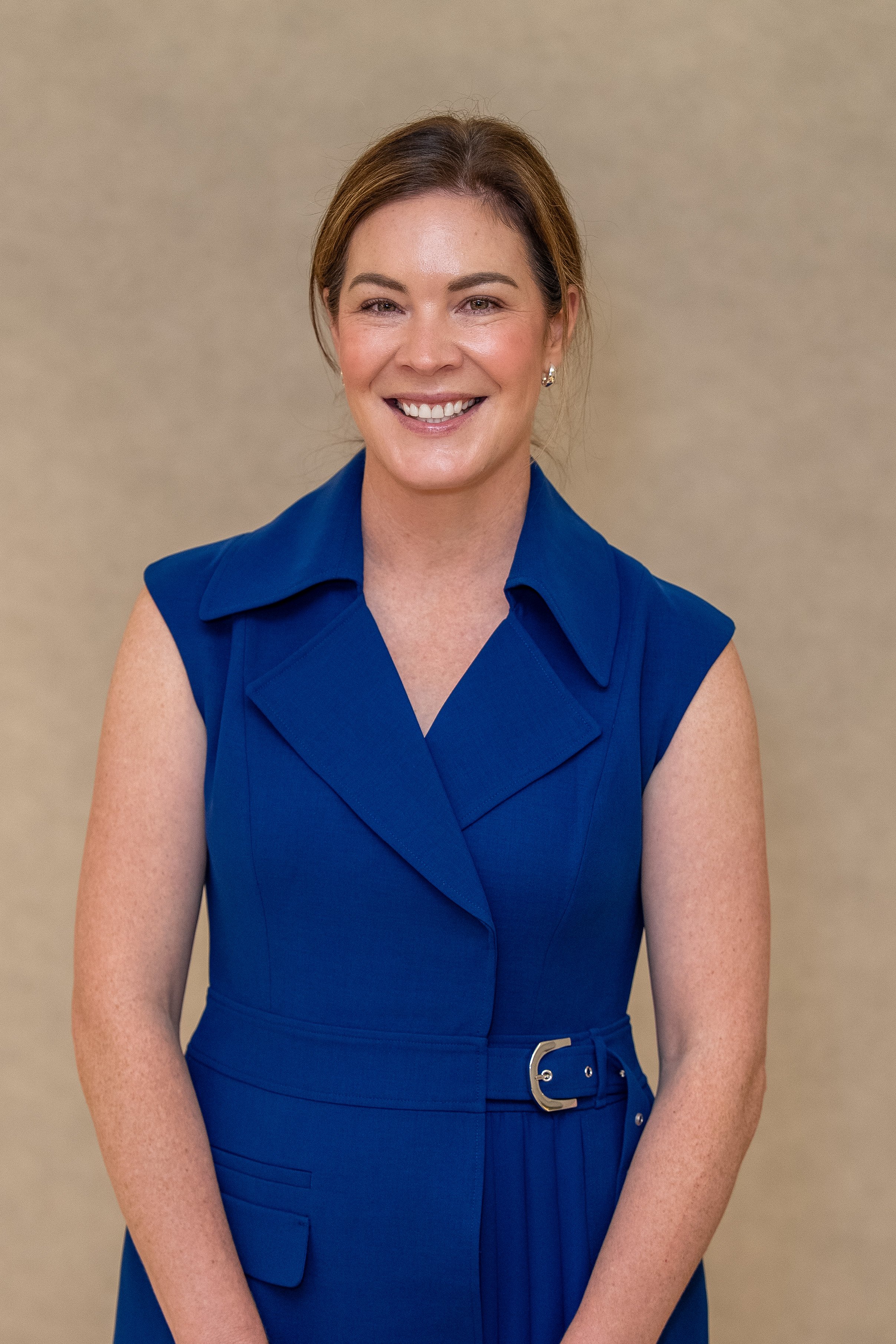 A woman with brown hair in a ponytail, wearing a sleeveless royal blue dress with a belt, smiling and standing against a beige background.