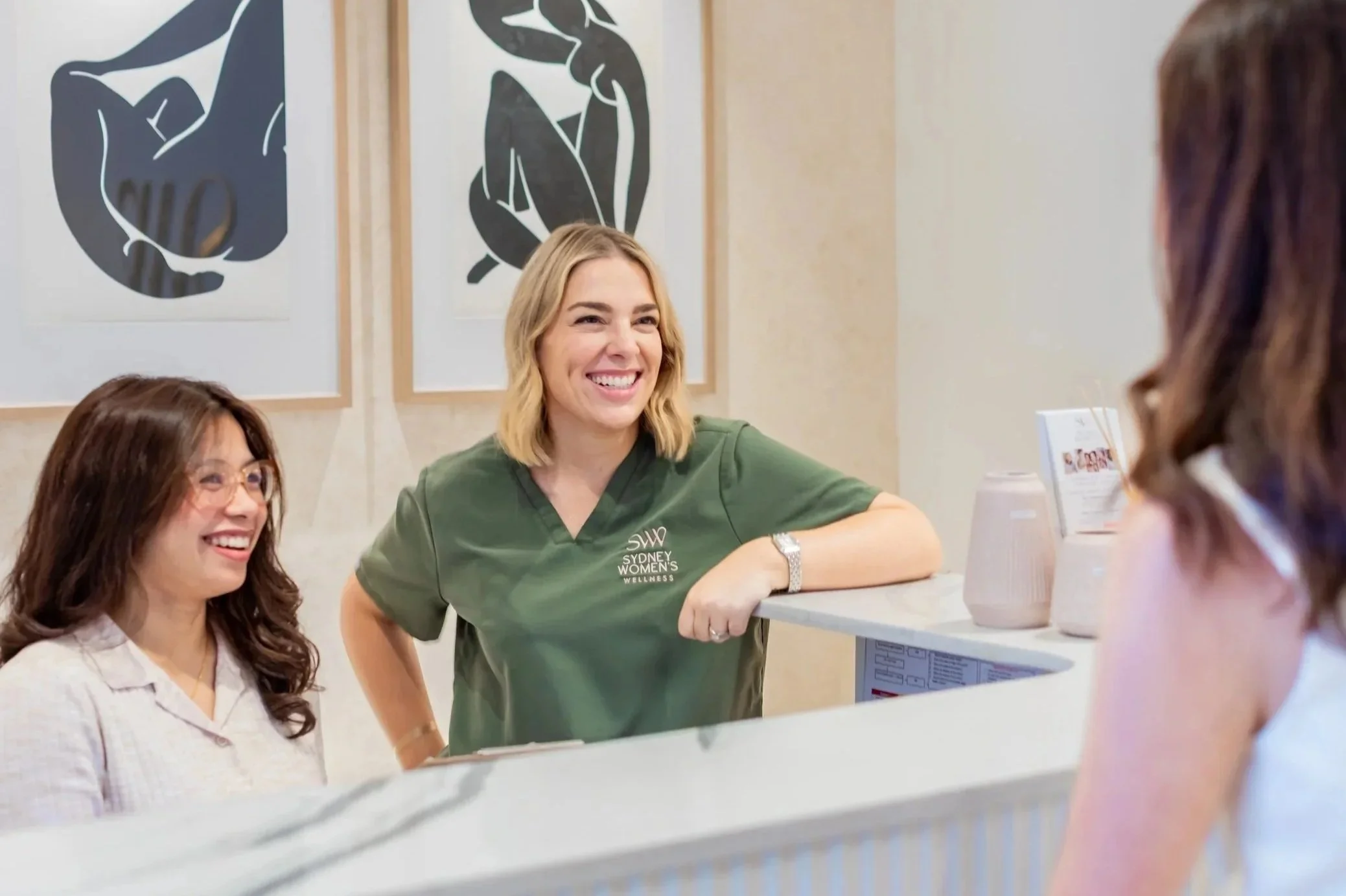 Group of women talking at a reception desk; one woman in green scrubs with 'Sydney Women's Wellness' embroidered, smiling among other women.