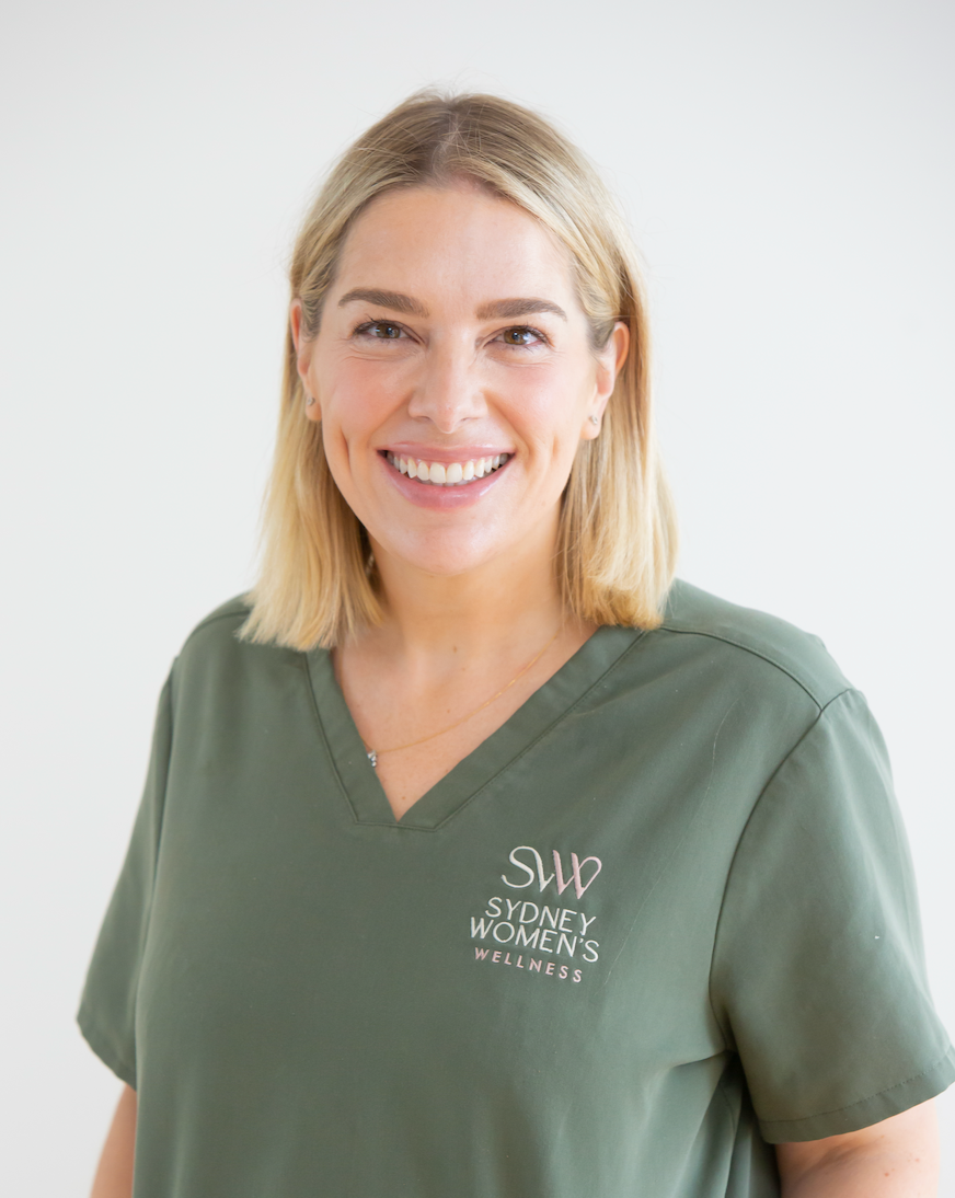 A woman smiling, wearing a green medical scrub top with the logo 'Sydney Women's Wellness' embroidered on it, standing against a plain white background.