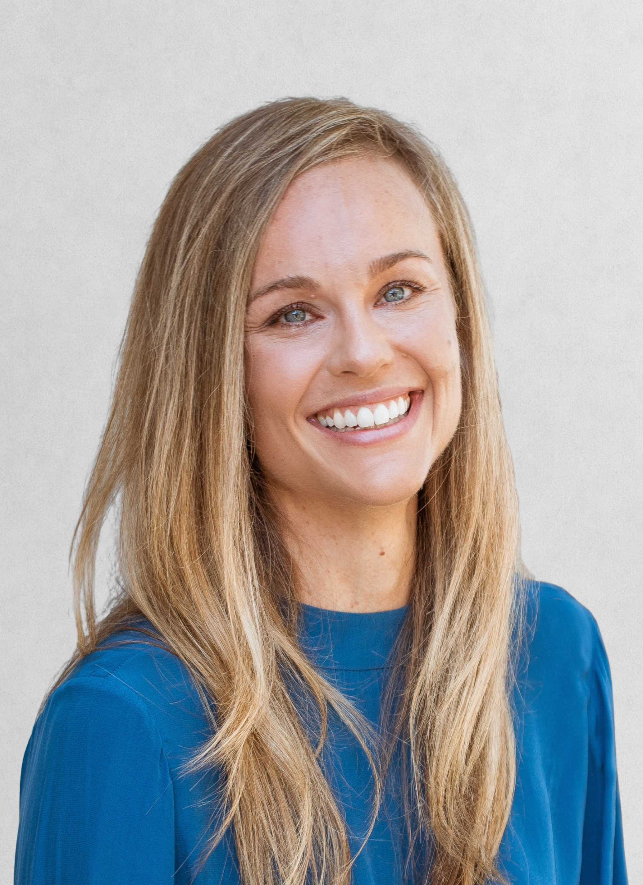 Close-up of a smiling woman with long blonde hair and blue eyes wearing a blue top against a plain light background.