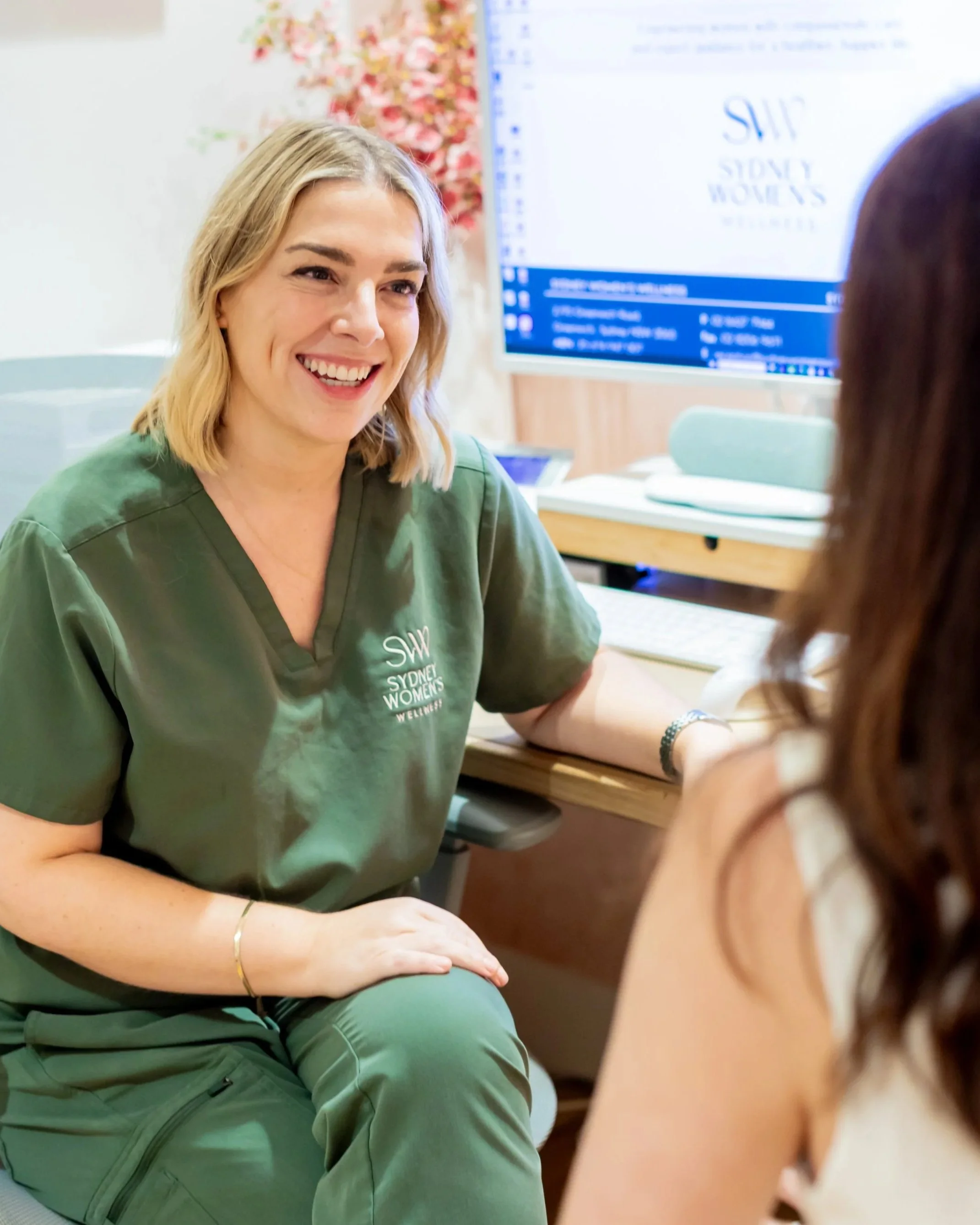 A woman with blonde hair smiling and talking to another woman with brown hair in an office setting. The woman in green scrubs has a laptop and a computer monitor in the background displaying the Sydney Women's group logo.