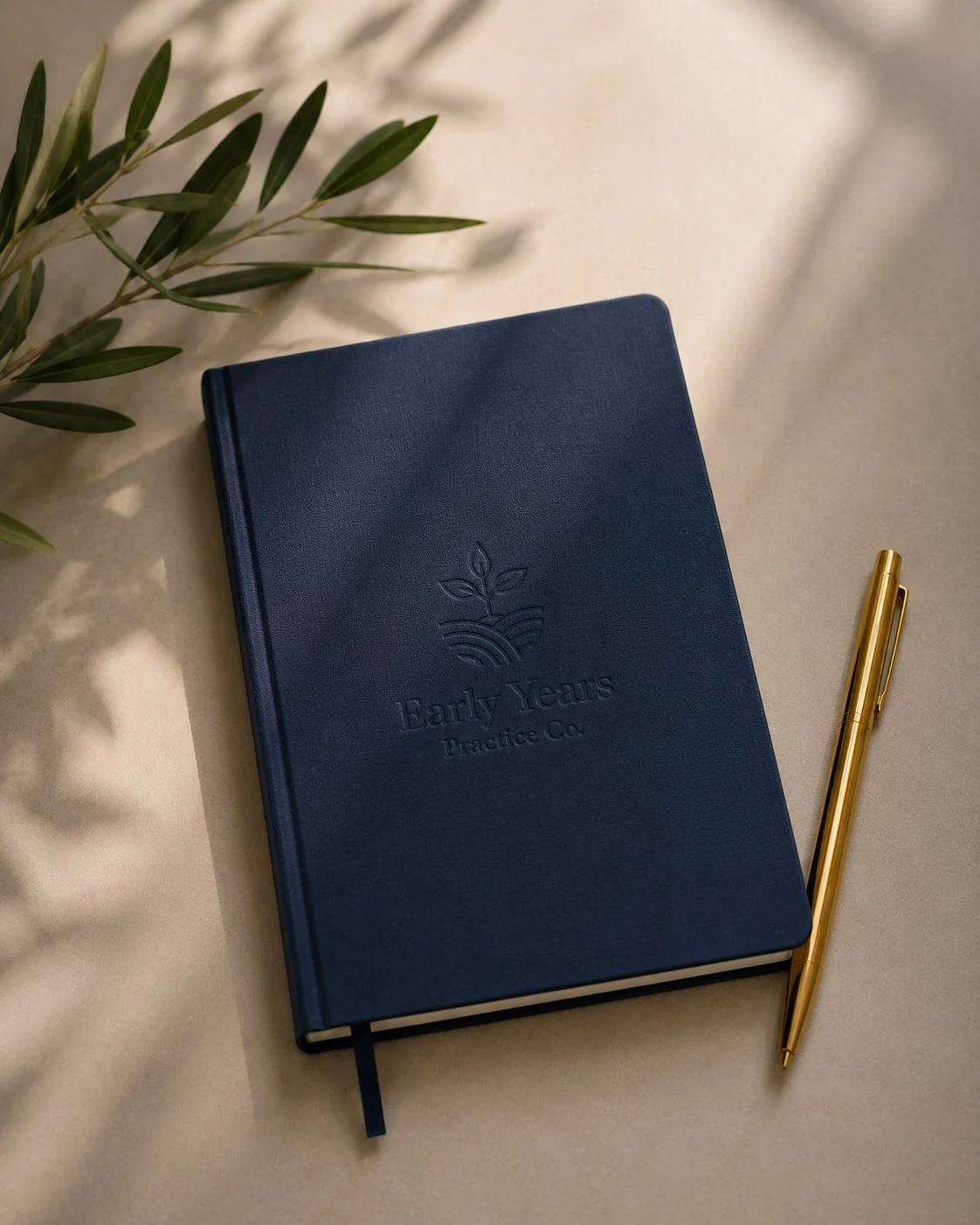 A blue hardcover notebook with the logo and text 'Early Years Practice Co.' embossed on the front, a gold pen lying next to it, and some green leaves partially visible in the upper left corner, all placed on a beige surface.