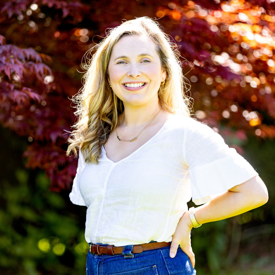 A woman with long blonde hair smiling outdoors, standing in front of red and green foliage, wearing a white top and blue jeans.