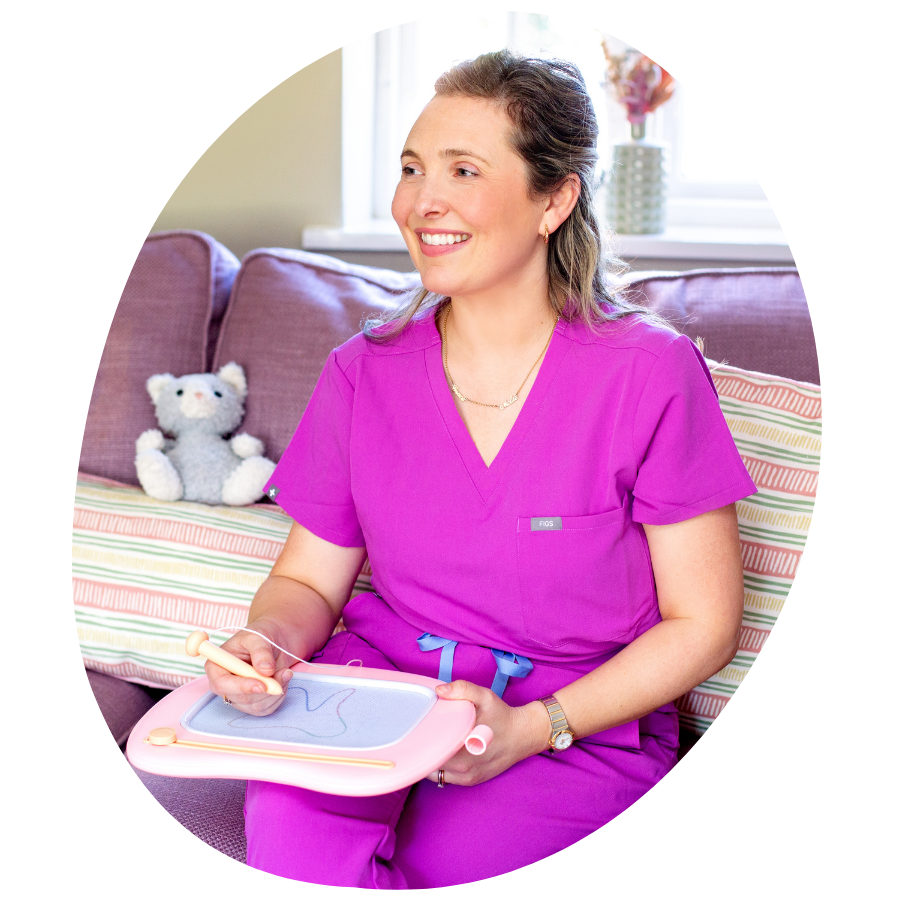 A woman in pink scrubs sitting on a sofa, holding a miniature embroidery or needlepoint frame, smiling with a teddy bear in the background.