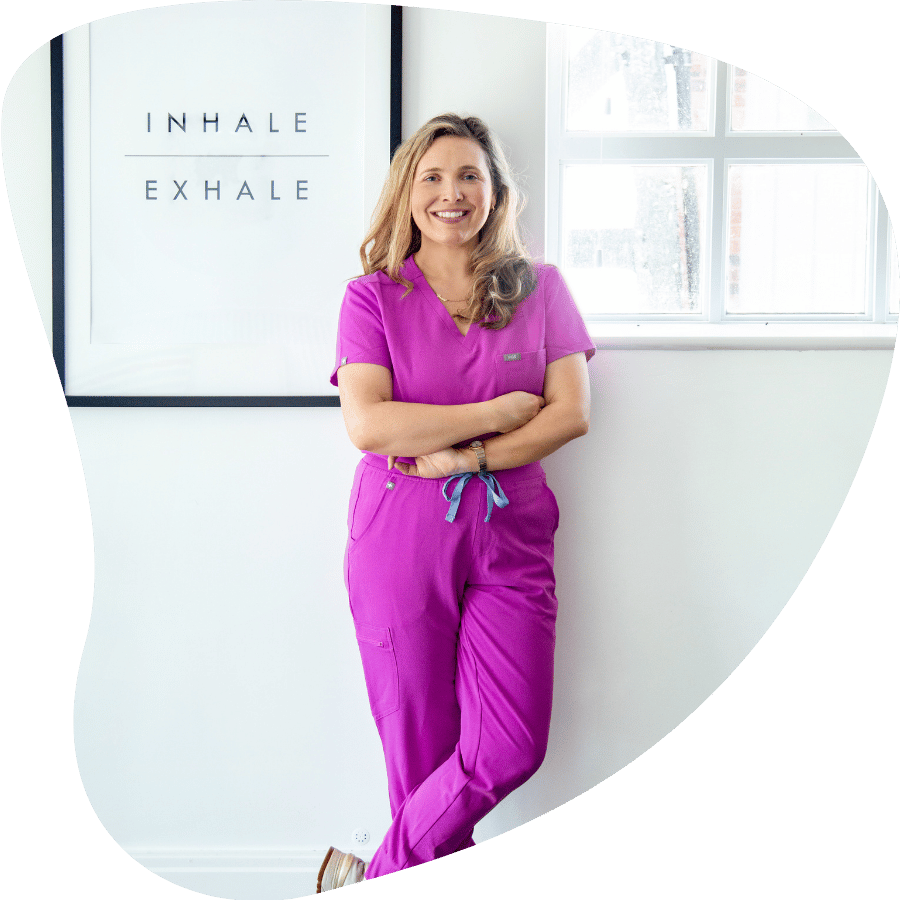 A smiling woman in pink scrubs standing in a medical office with her arms crossed, in front of a white wall with a sign that says 'Inhale' and 'Exhale' and a window.