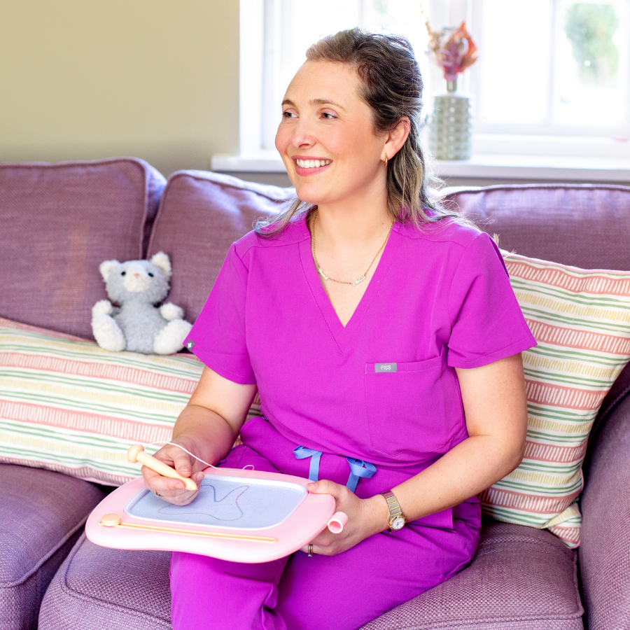 A smiling woman in pink scrubs sitting on a purple couch with a teddy bear and striped pillows, holding a drawing board and stylus in a bright room.