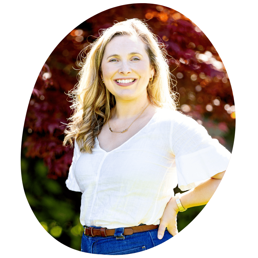 A woman with blonde hair smiling outdoors in front of red autumn trees, wearing a white blouse, blue jeans, and a gold watch.