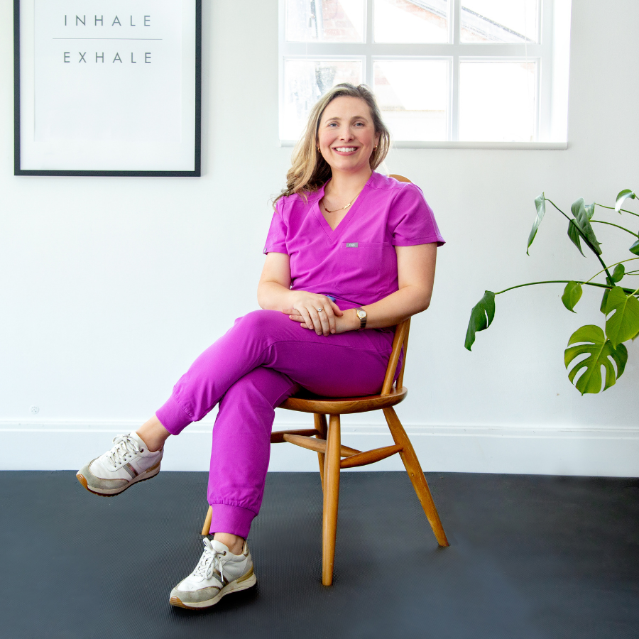 A woman in purple scrubs sitting on a wooden chair with her legs crossed, smiling, in a bright room with a window, green plant, and motivational poster that says "Inhale" and "Exhale."
