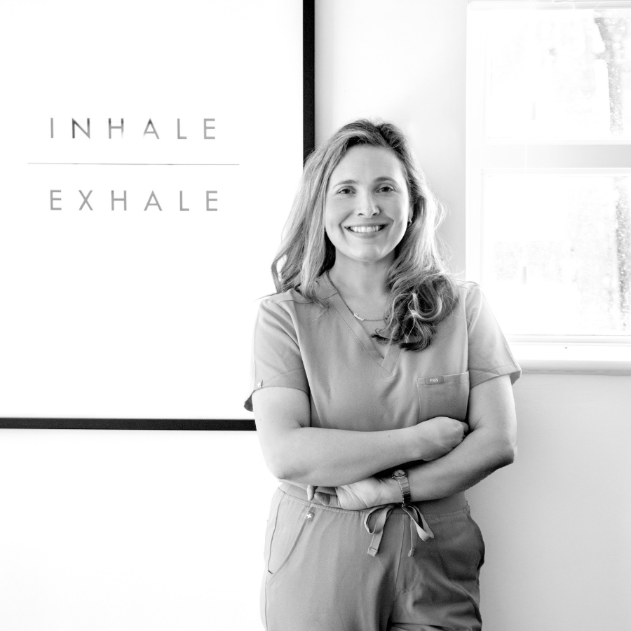 A woman with long, wavy hair smiling and standing with her arms crossed next to a whiteboard that says 'Inhale Exhale' in a bright, sunlit room.