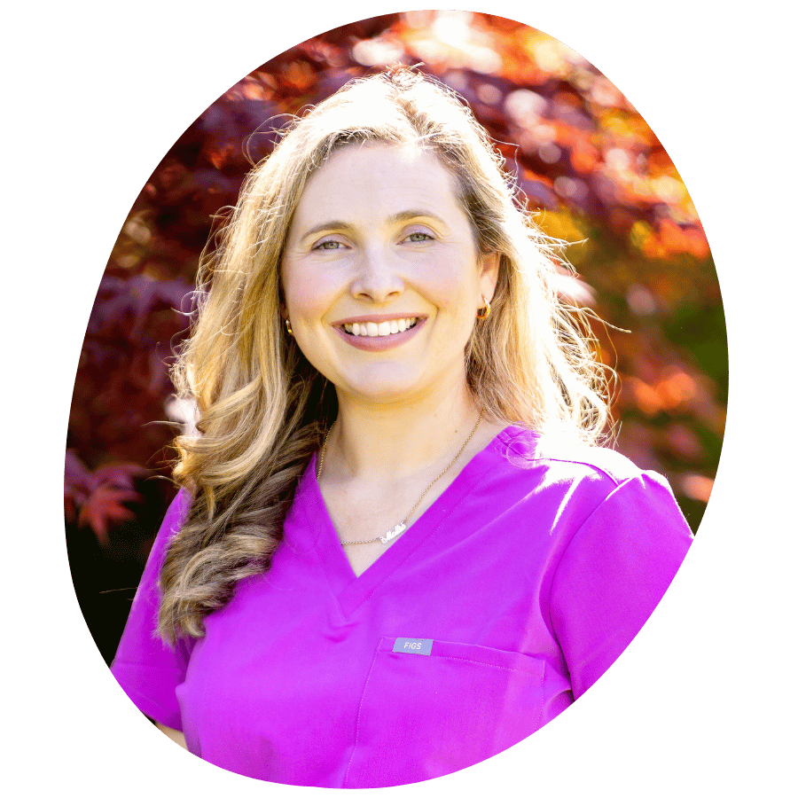 A woman with long blonde hair smiling, wearing purple scrubs, standing outdoors with colorful autumn leaves in the background.