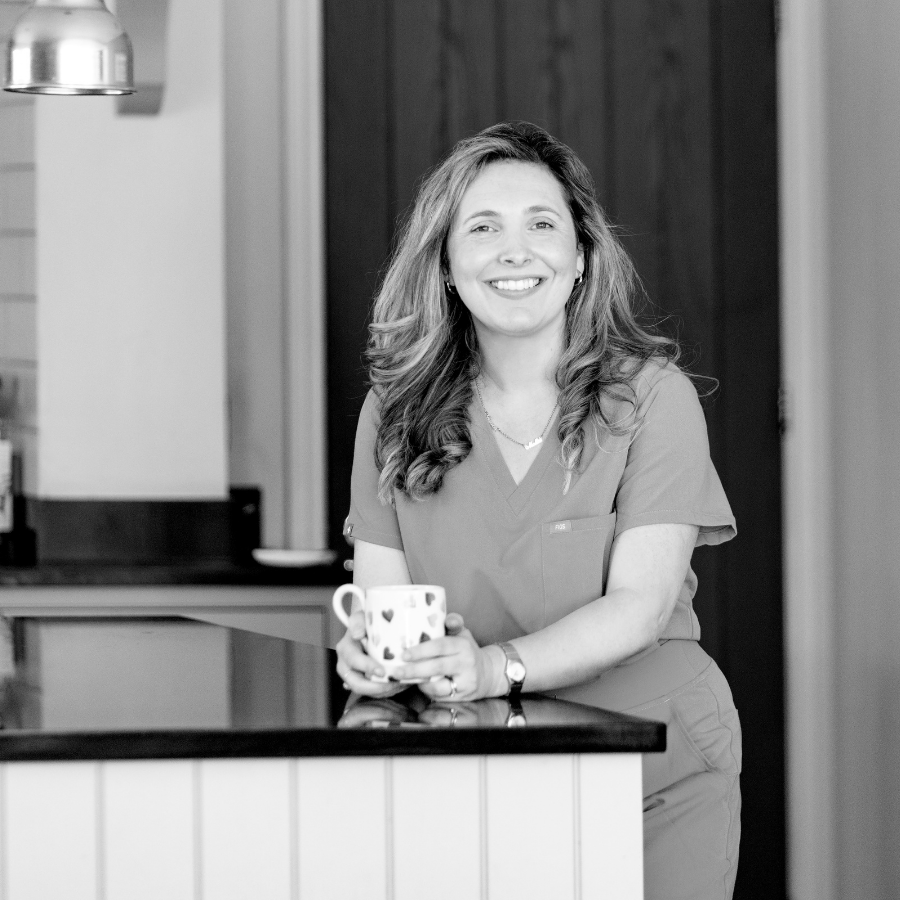 A smiling woman with long hair wearing medical scrubs, holding a mug, standing in a kitchen or break room setting.