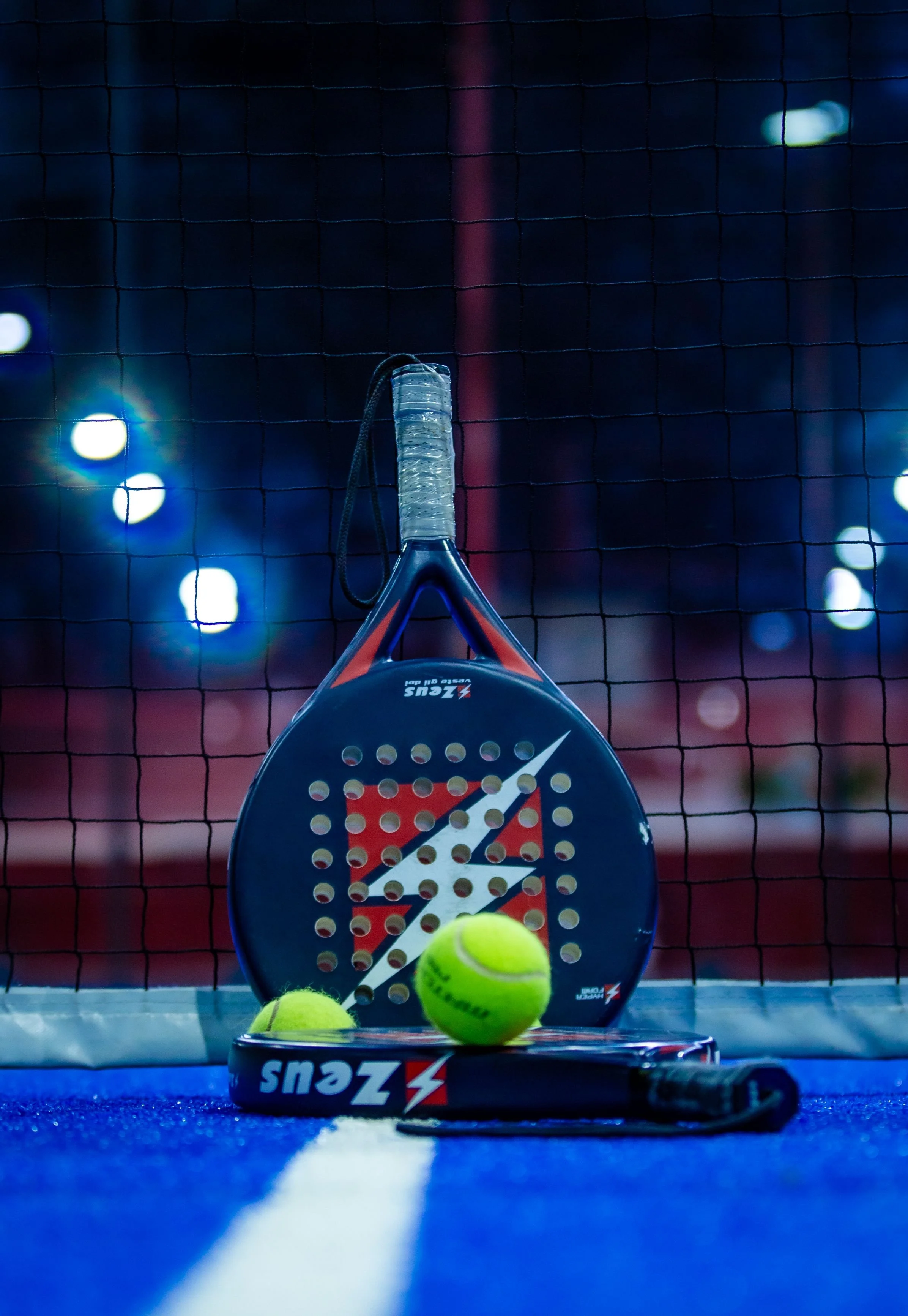 Tennis rackets and tennis balls on a court, with a net in the background and blurred stadium lights, indicating an indoor tennis court setting.