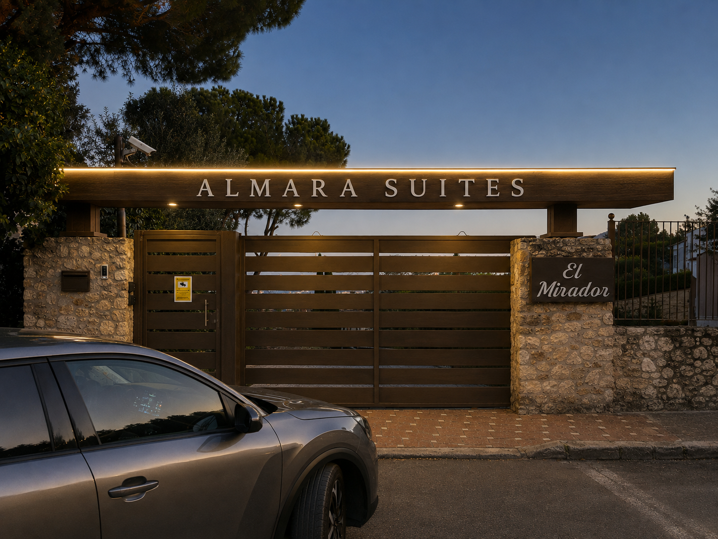 Entrance gate of Almará Suites, with stone columns, wooden fencing, and illuminated signage. A silver car is parked in front. Sign on right reads 'El Mirador'.