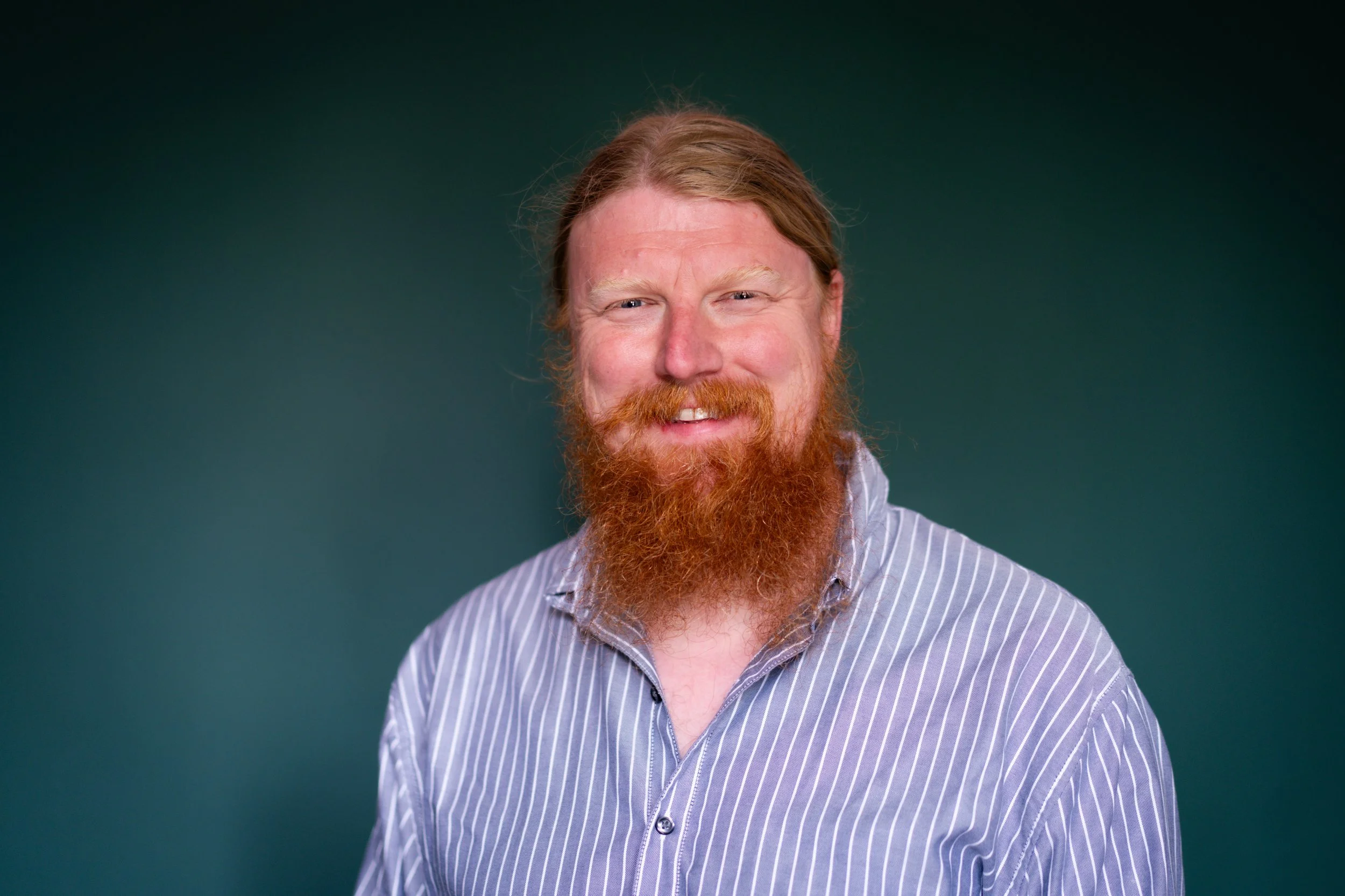 A man with long red hair and a full red beard smiling, wearing a light purple and white striped shirt, against a dark green background.