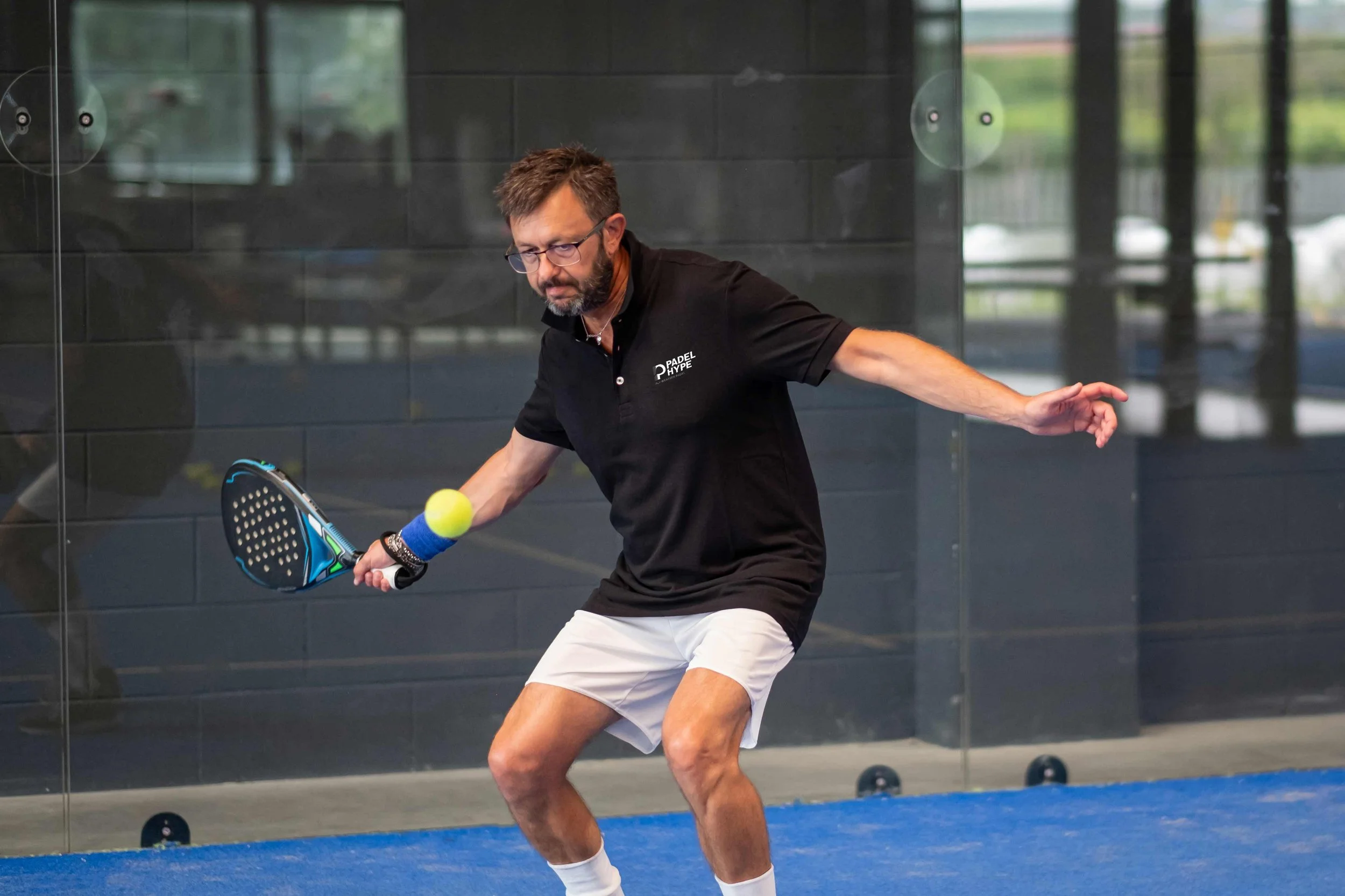 A man playing paddle tennis indoors, wearing a black polo shirt and white shorts, hitting a yellow tennis ball with a paddle, with a black wall and glass windows in the background.