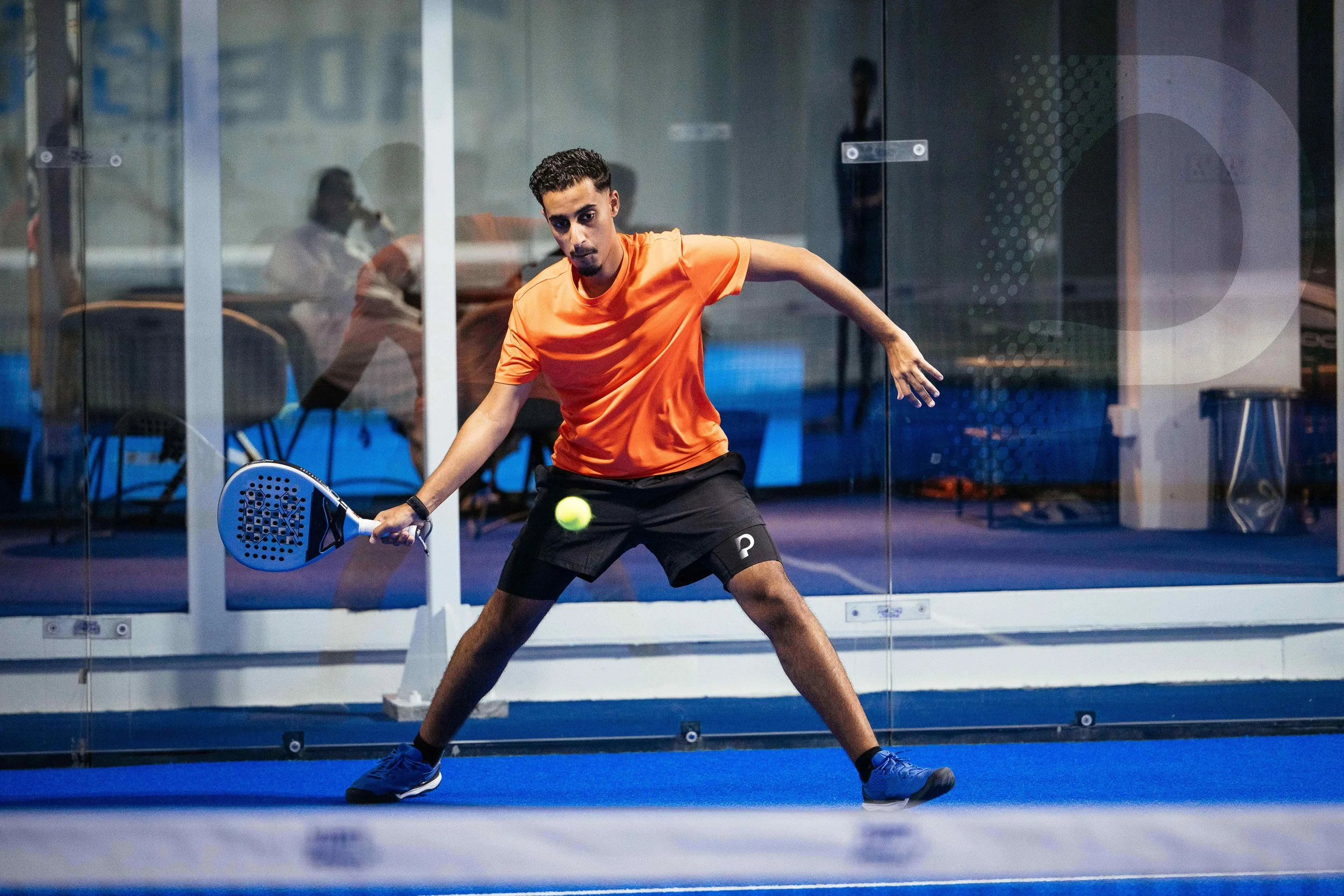 A young man in an orange shirt and black shorts is playing pickleball indoors, about to hit a yellow ball with a paddle while standing on a blue court behind a glass wall.