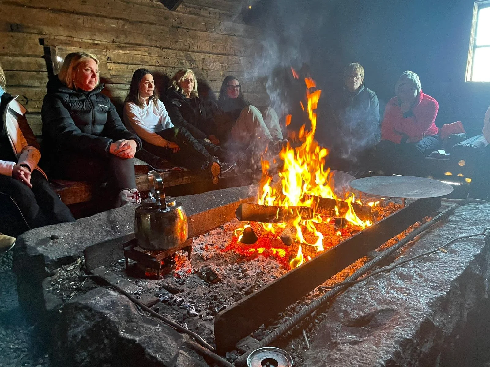 Gruppo di persone sedute intorno a un camino aperto in una stanza di legno, con il fuoco acceso al centro.