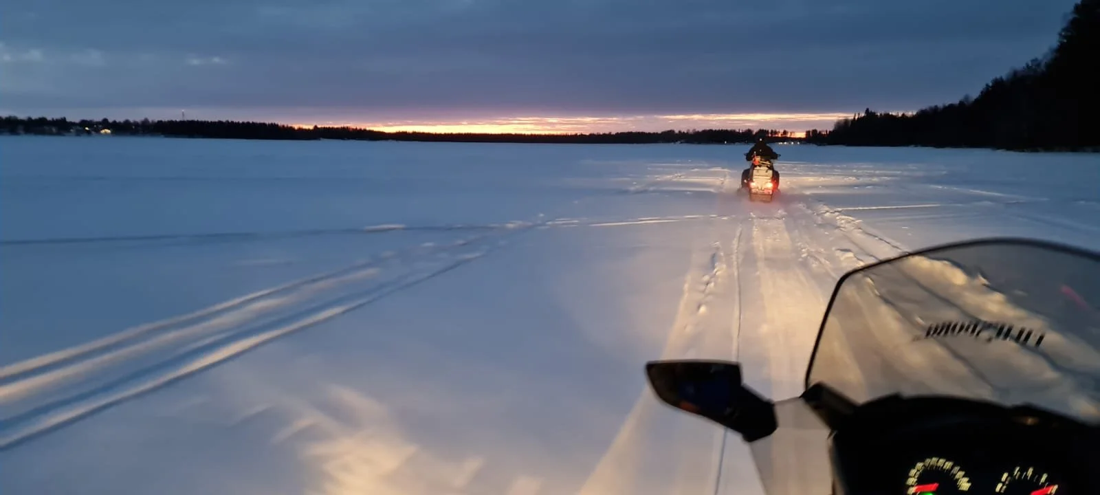 Due motoslitte che viaggiano su un campo di neve durante il tramonto, con il cielo nuvoloso e l'orizzonte illuminato da una sottile linea di luce
