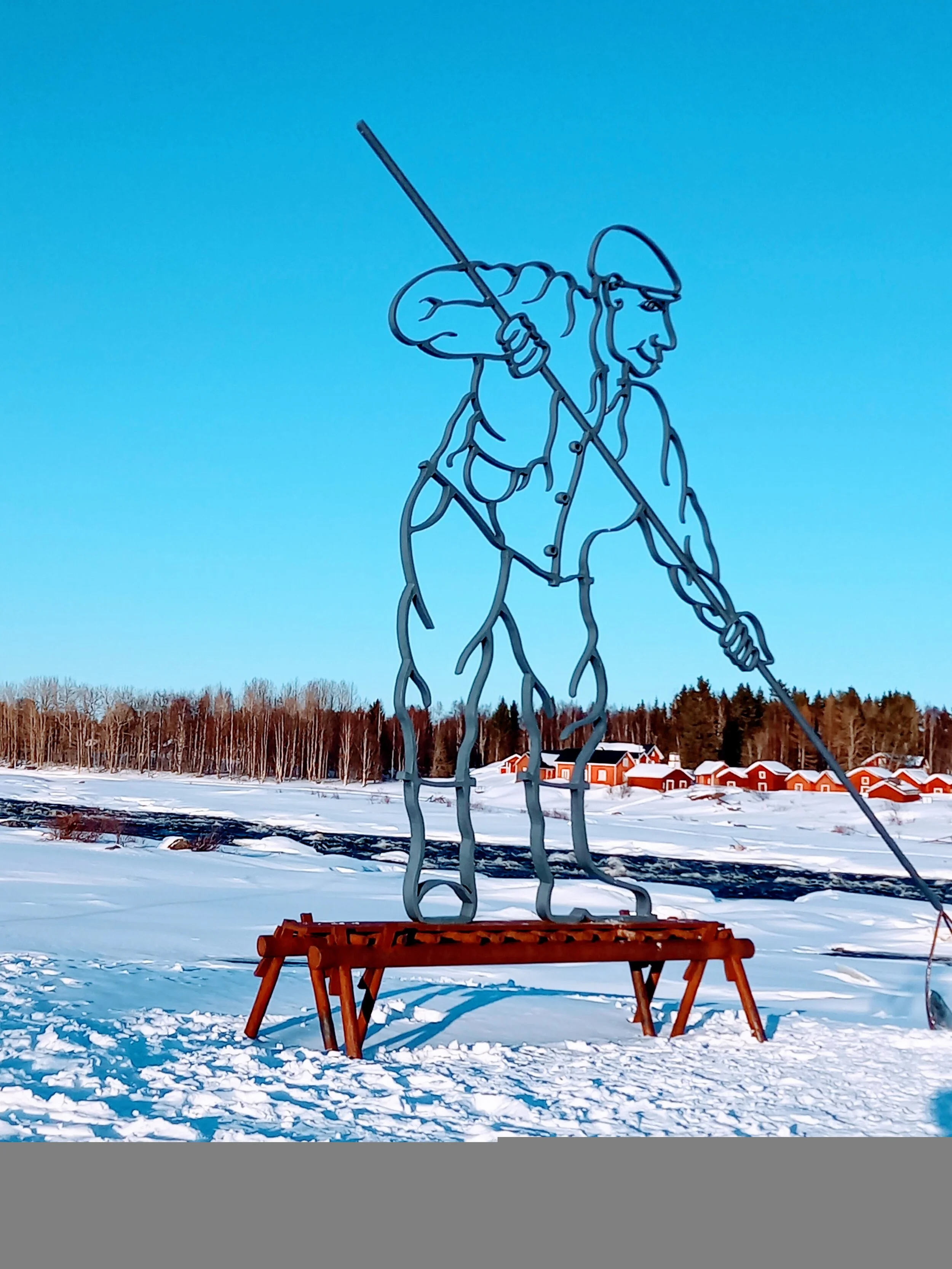 Scultura di un pescatore in metallo con un cappello, che tiene un'asta da pesca, in un paesaggio invernale con neve, alberi e case rosse sullo sfondo.