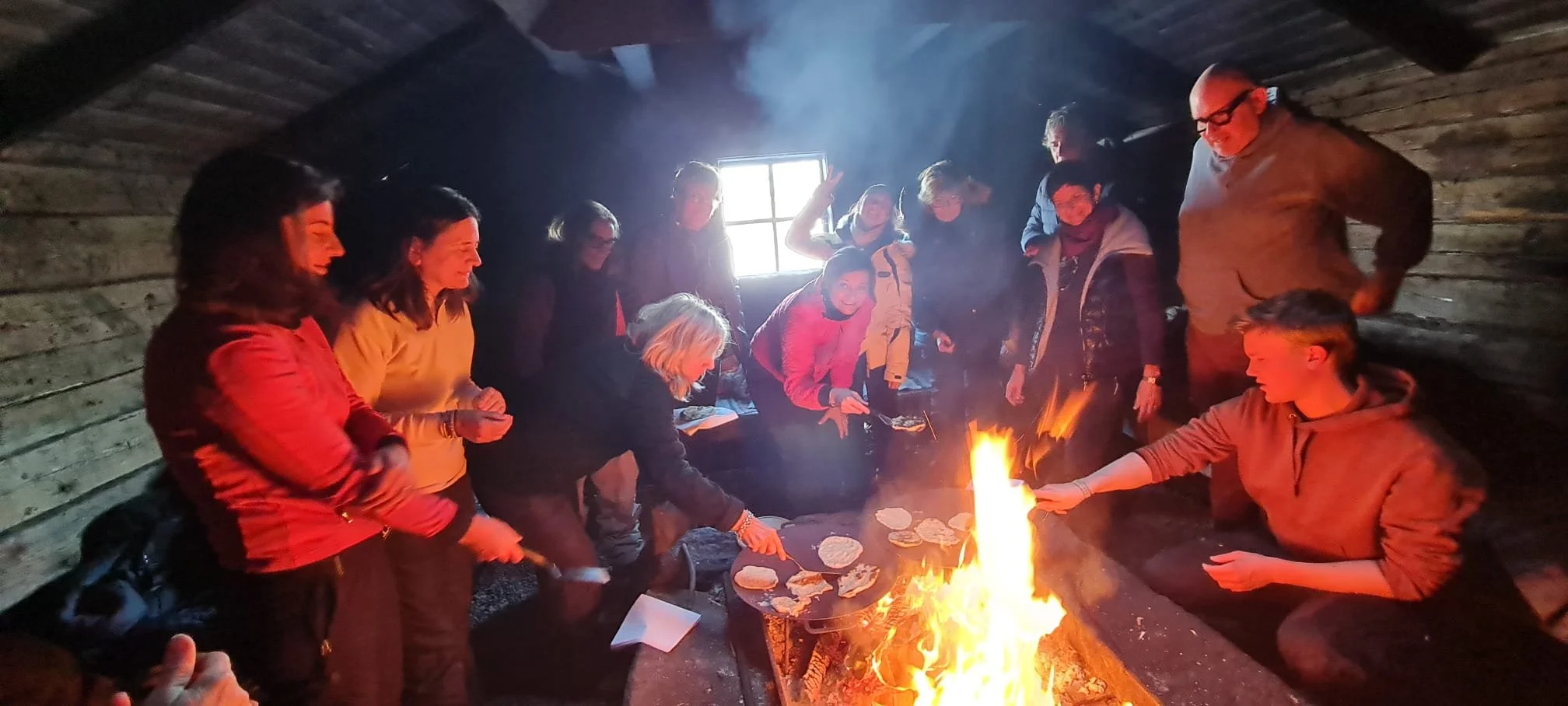 Gruppo di persone riunite intorno a un falò in una stanza di legno, alcune stanno cucinando o bruciando pane, sorridendo insieme.