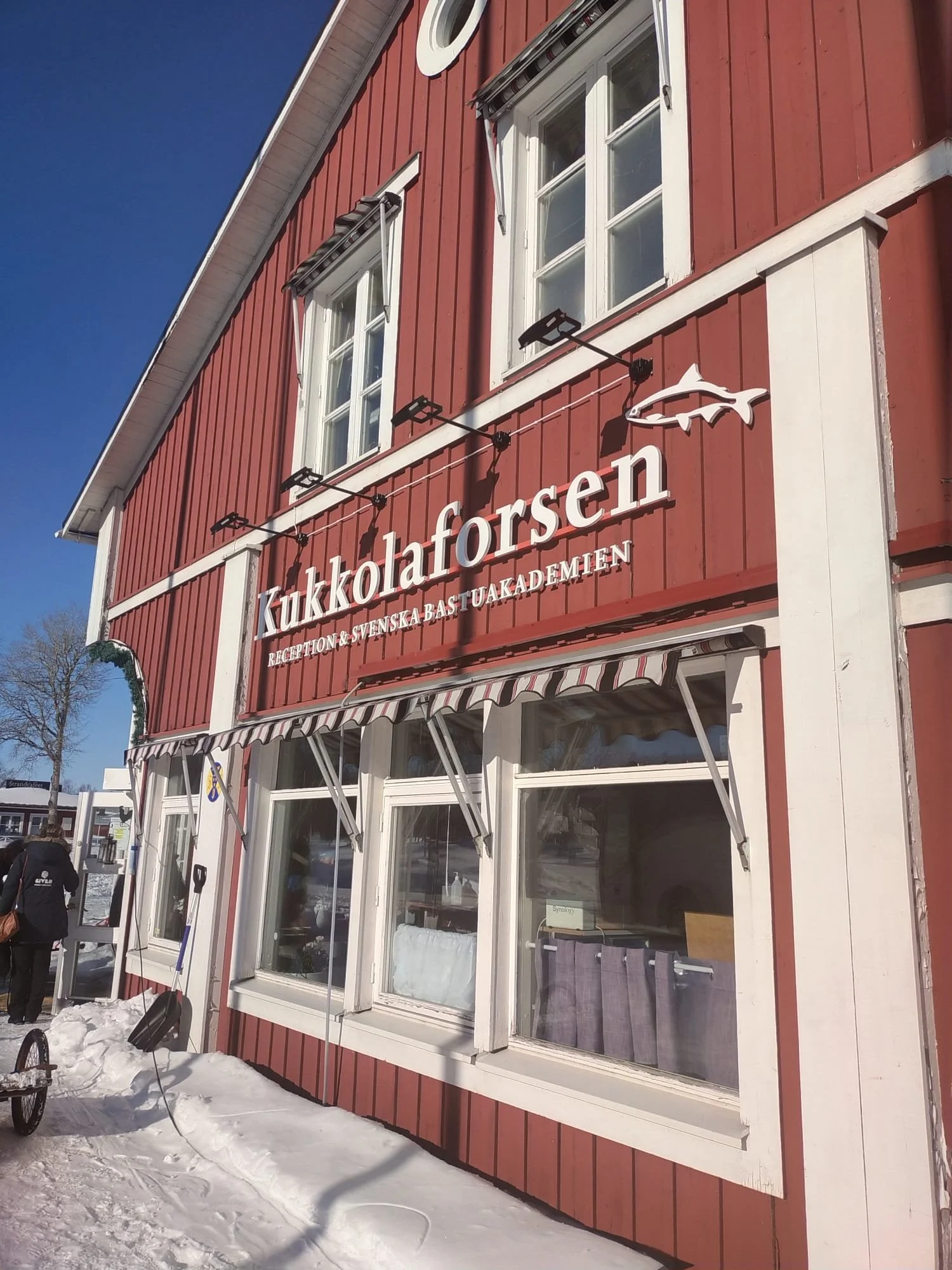 Esterno di un edificio rosso con finestre bianche e insegna con parola "Kukkolaforsen" e simbolo di un pesce, sotto scritto "Reception & Svenska Bastu Akademien".