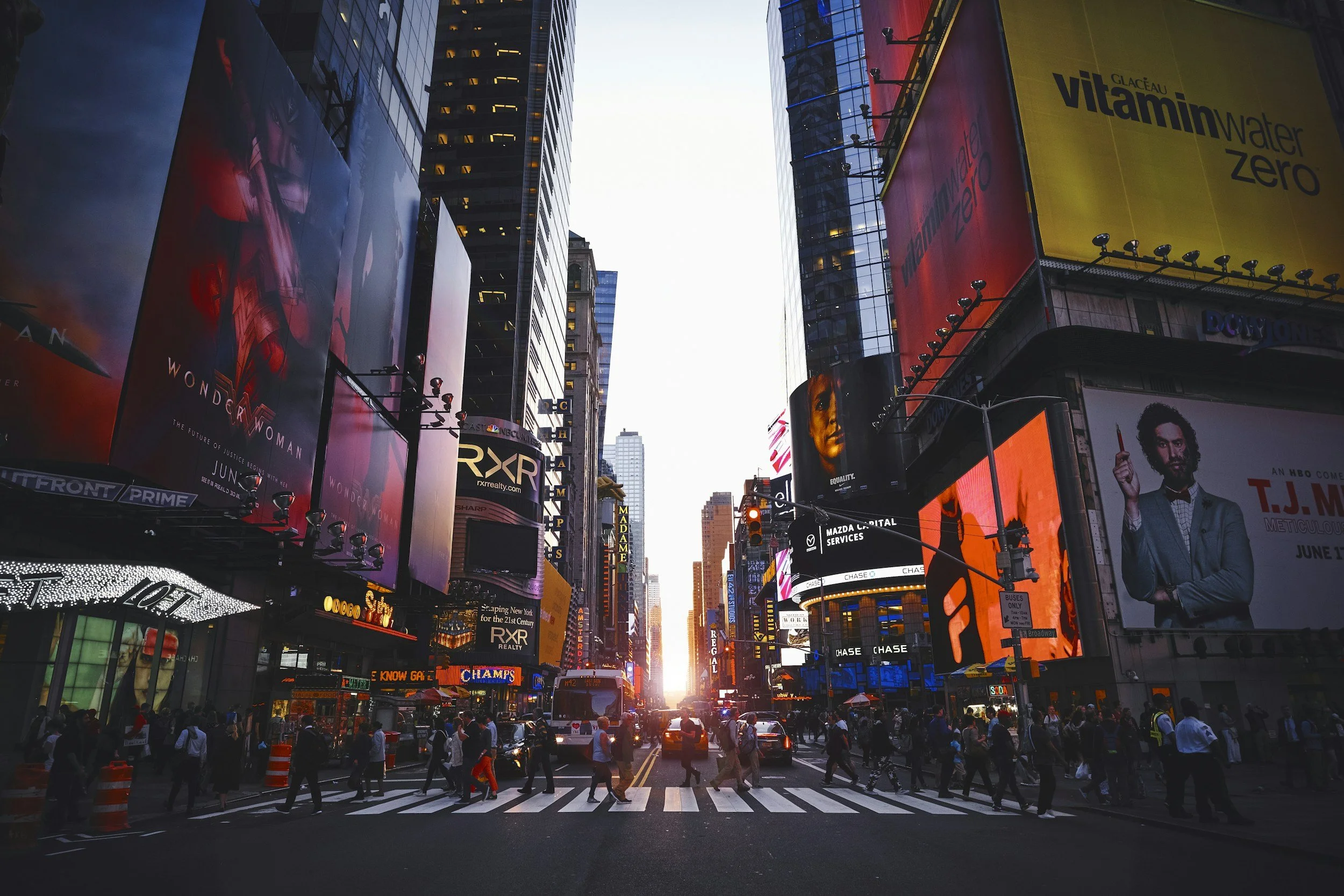 Una scena di Times Square a New York con grandi cartelloni pubblicitari illuminati, traffico e persone che attraversano la strada al tramonto.