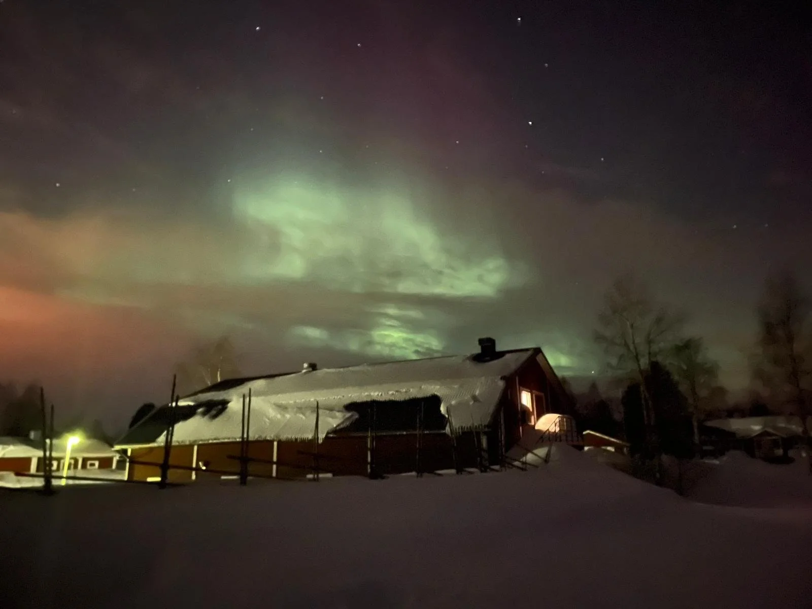 Notte con aurora boreale verde visibile nel cielo sopra una casa con tetto innevato e alberi nudi, atmosfera invernale.