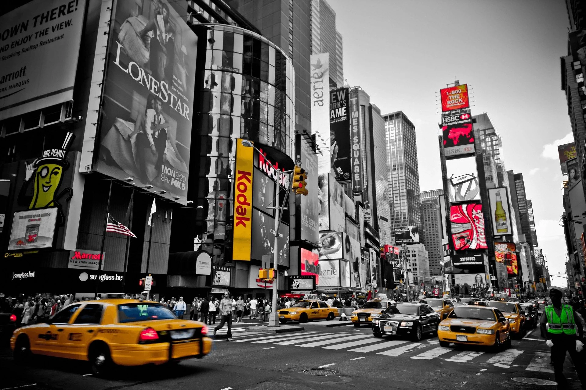 Immagine in bianco e nero di Times Square a New York con cartelloni pubblicitari, macchine gialle, pedoni, e un addetto alla sicurezza con giacca verde.