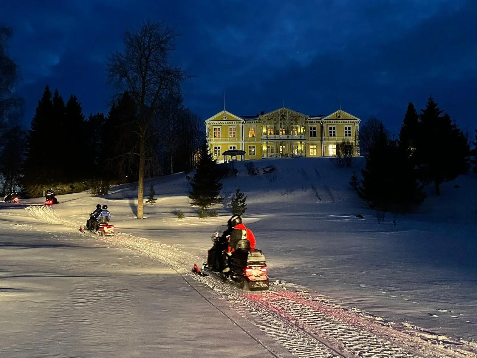 Escursione in motoslitta sulla neve davanti a una grande villa illuminata di notte, con un cielo scuro e alberi intorno.