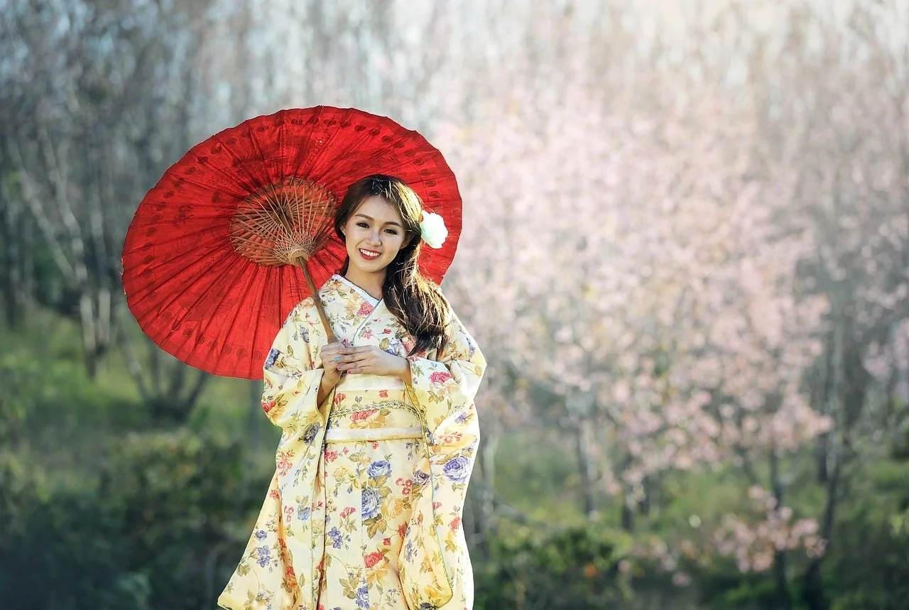 Una donna che indossa un kimono giallo con fiori, sorride e tiene un ombrello rosso in un paesaggio con alberi in fiore.