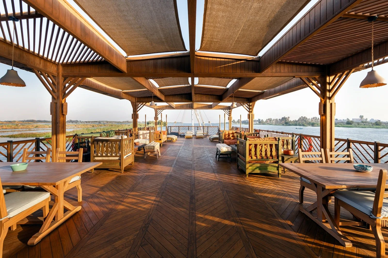 Terrazza in legno con vista sul fiume, arredamento con sedie, tavoli, divani in stile rustico, ampie coperture di tessuto e luci da soffitto, al tramonto.