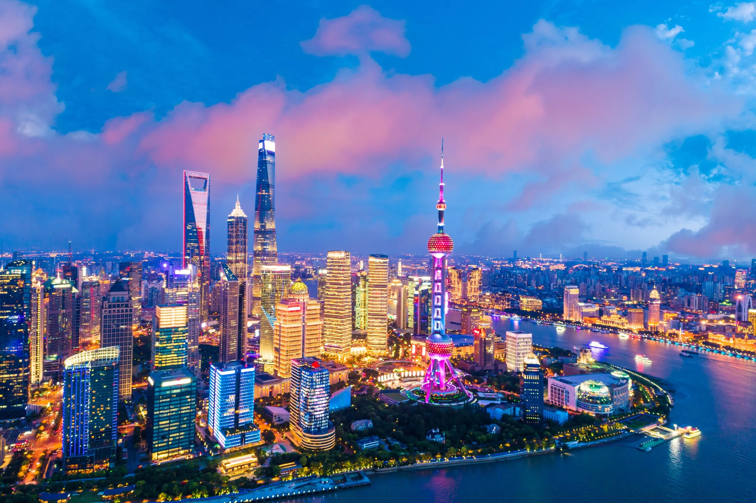 Skyline di Shanghai di sera con grattacieli illuminati, tra cui la Torre delle Perle e la Shanghai World Financial Center, con cielo blu e nuvole rosa sopra la città