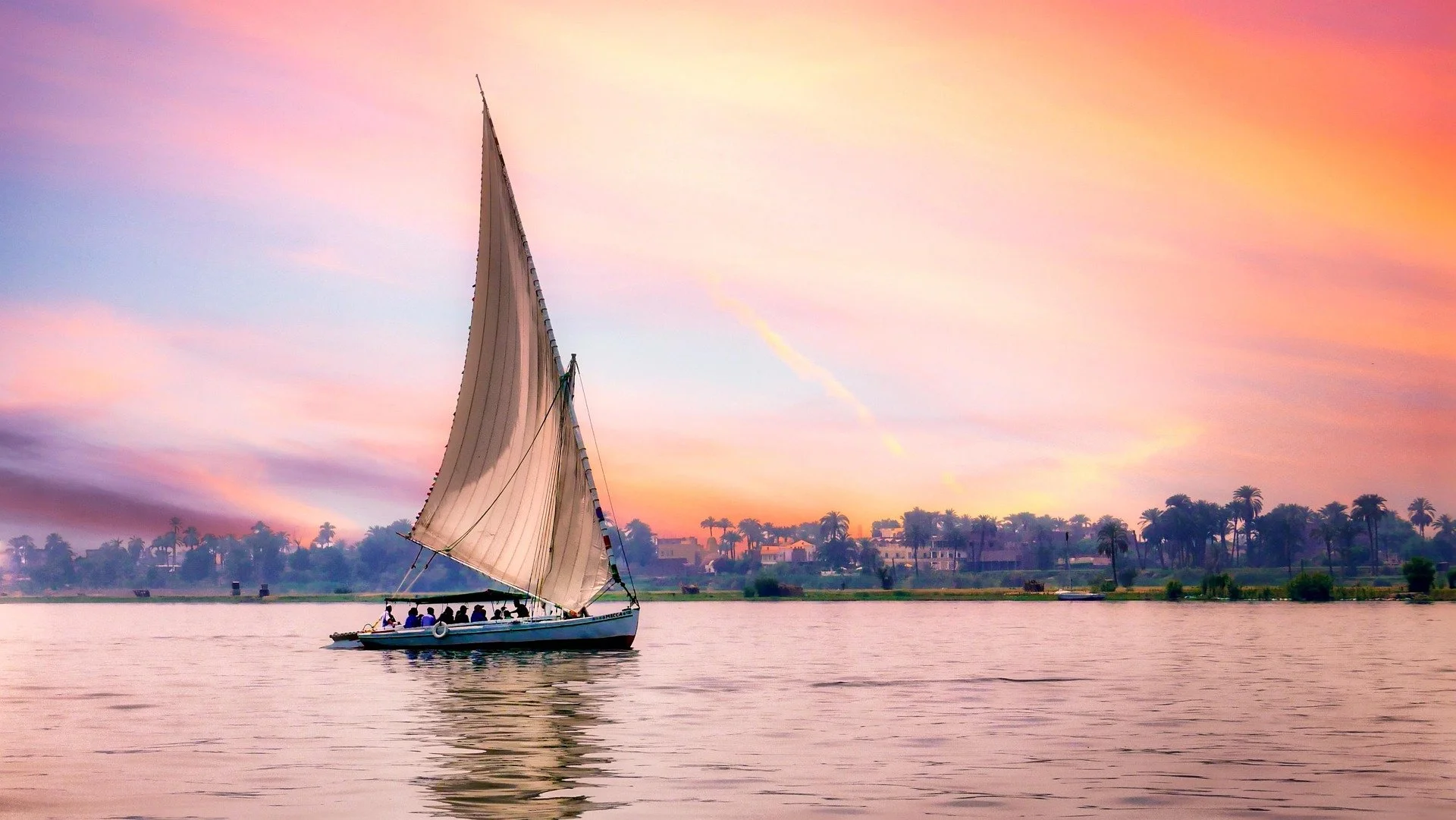 Una barca a vela naviga su un fiume durante un tramonto con il cielo dipinto di colori rosa, arancioni e viola, con un paesaggio di palme e case sullo sfondo.