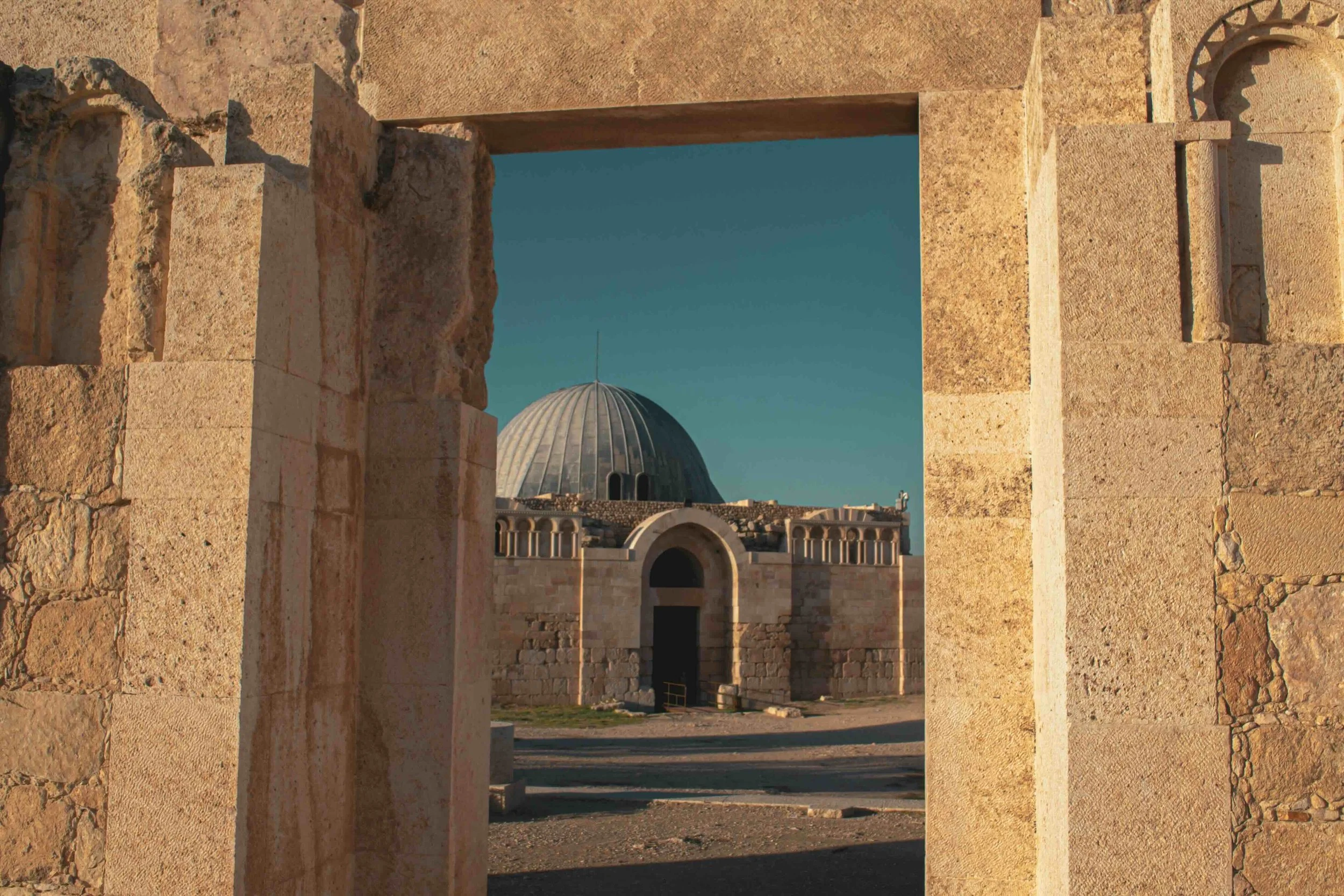 Resti di un'antica struttura con arcate e un grande tetto a cupola, visibile attraverso un'apertura architettonica in un muro di pietra.