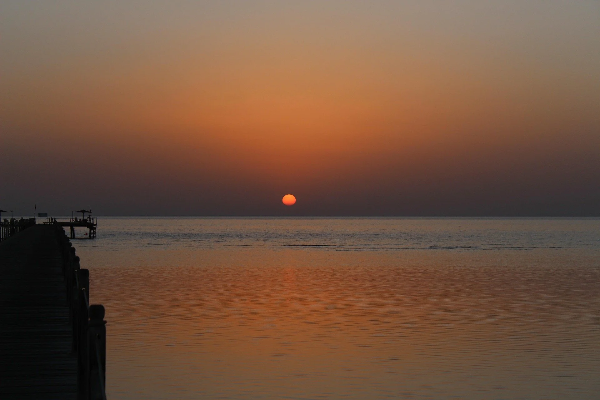 Tramonto sul mare visto da una banchina, con il sole arancione all'orizzonte e cielo sfumato