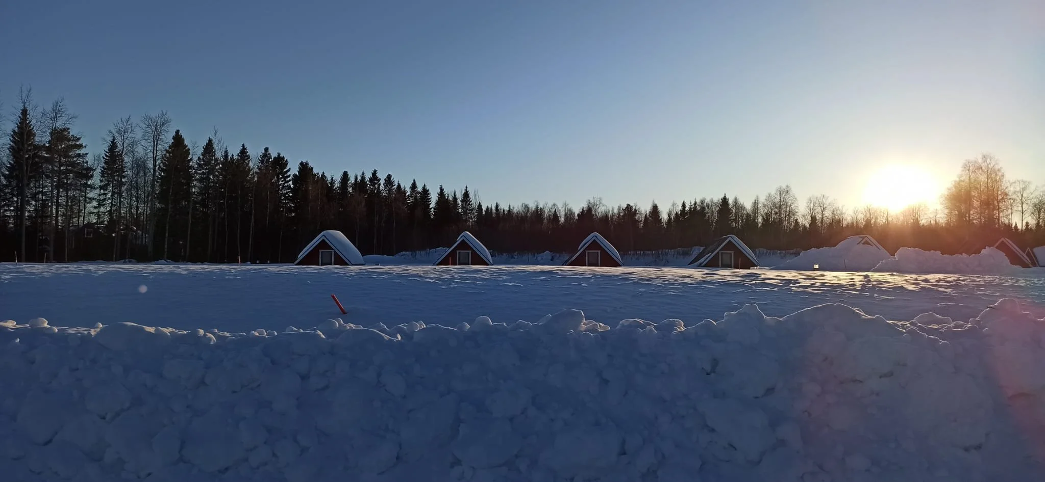 Case di legno in stile A con tetti ricoperti di neve in un paesaggio invernale, con alberi e il sole che tramonta all'orizzonte.