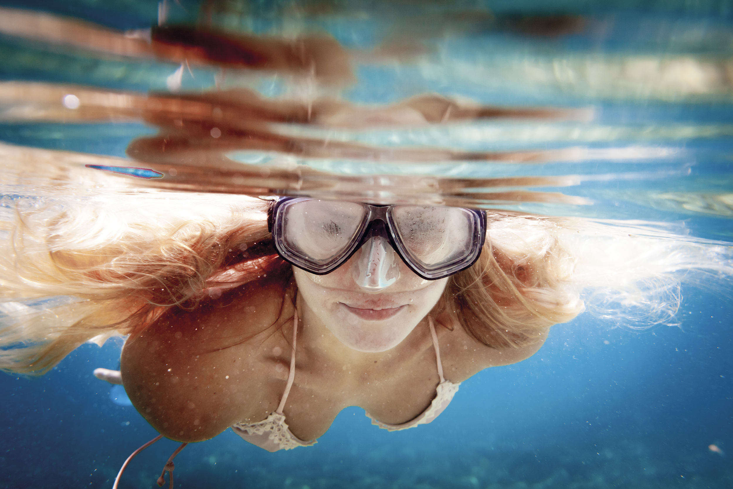 Una donna con capelli lunghi e chiari indossa occhialini da nuoto ed è sott'acqua in piscina, sorridendo mentre nuota.