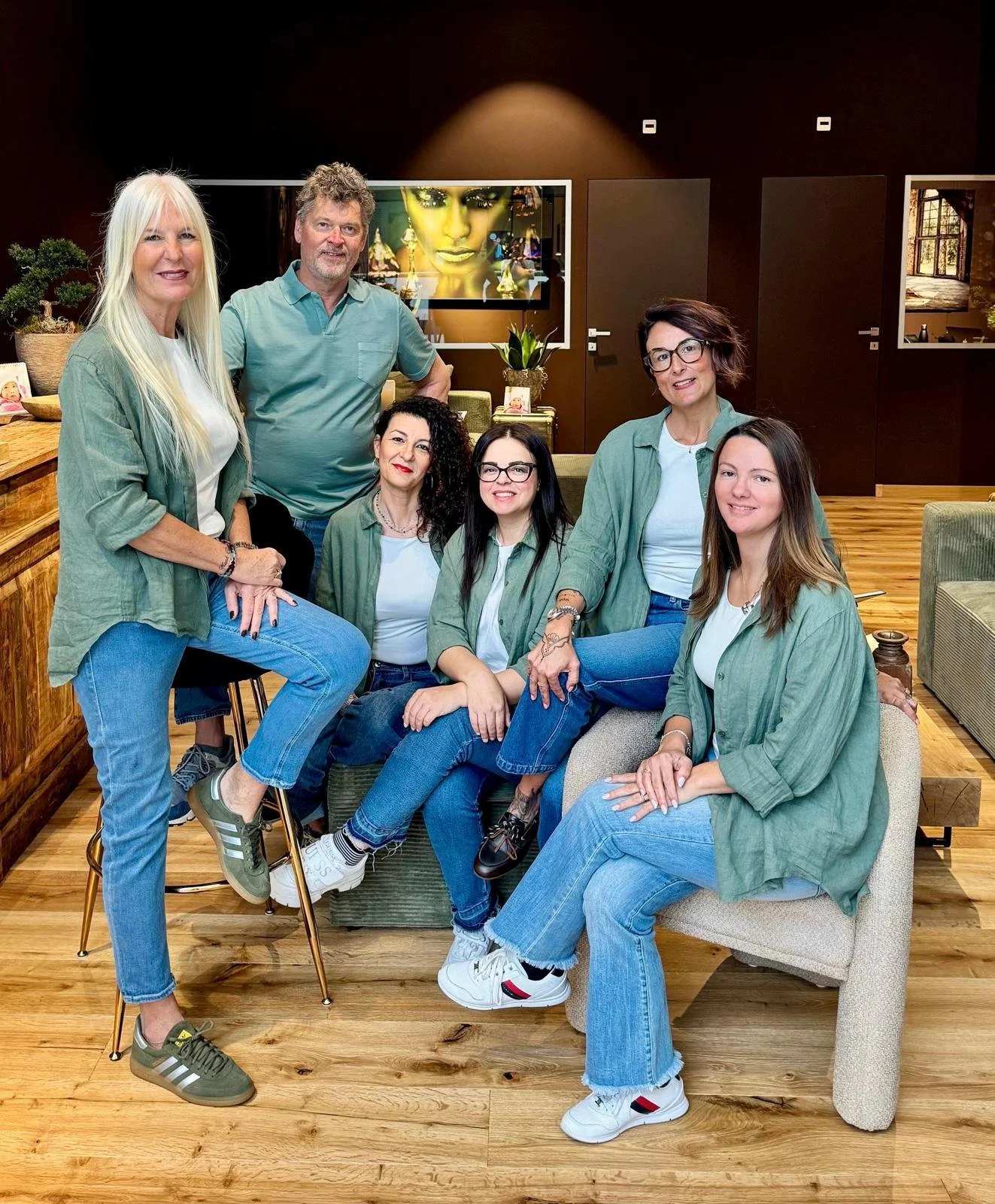 Gruppo di sei donne e un uomo seduti e in piedi in un ambiente domestico, con pareti marroni e fotografie alle pareti, tutti indossano abiti casual con camicie verdi o bianche e jeans.