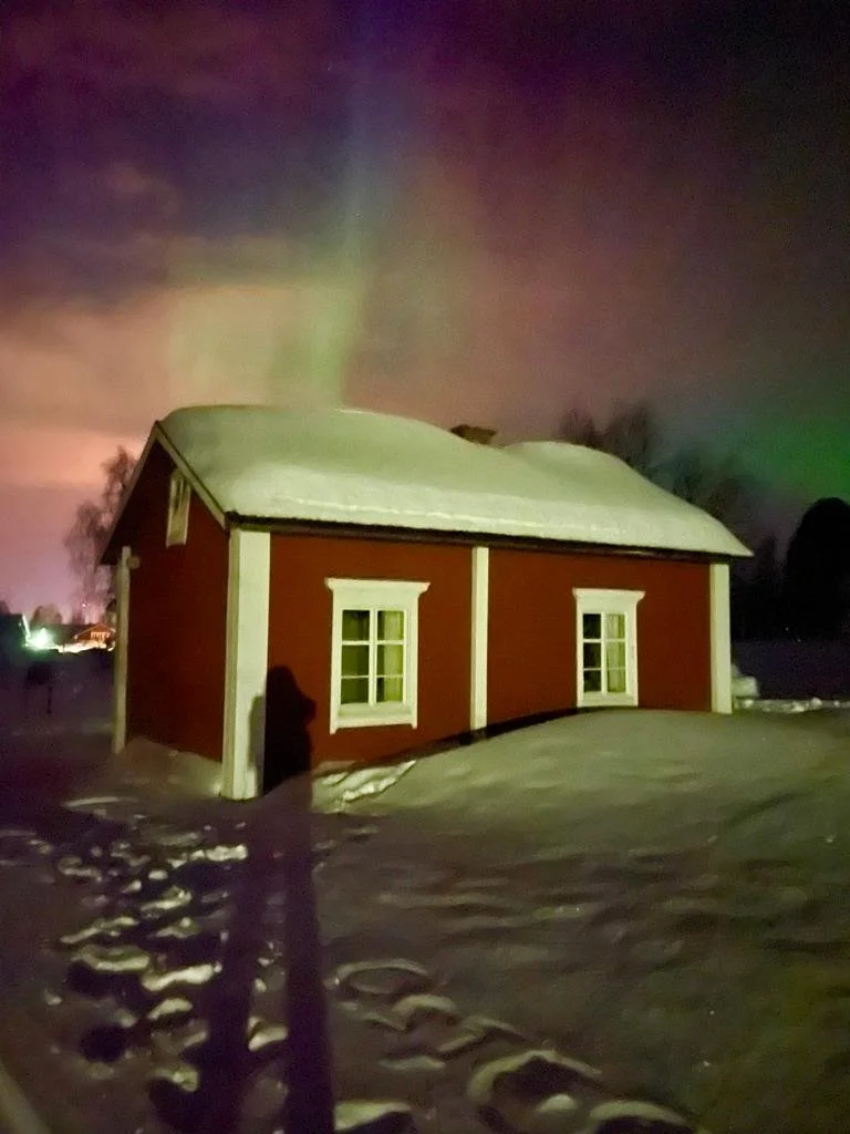 Una casetta di legno rossa con infissi bianchi coperta di neve sotto un cielo notturno con aurore boreali.